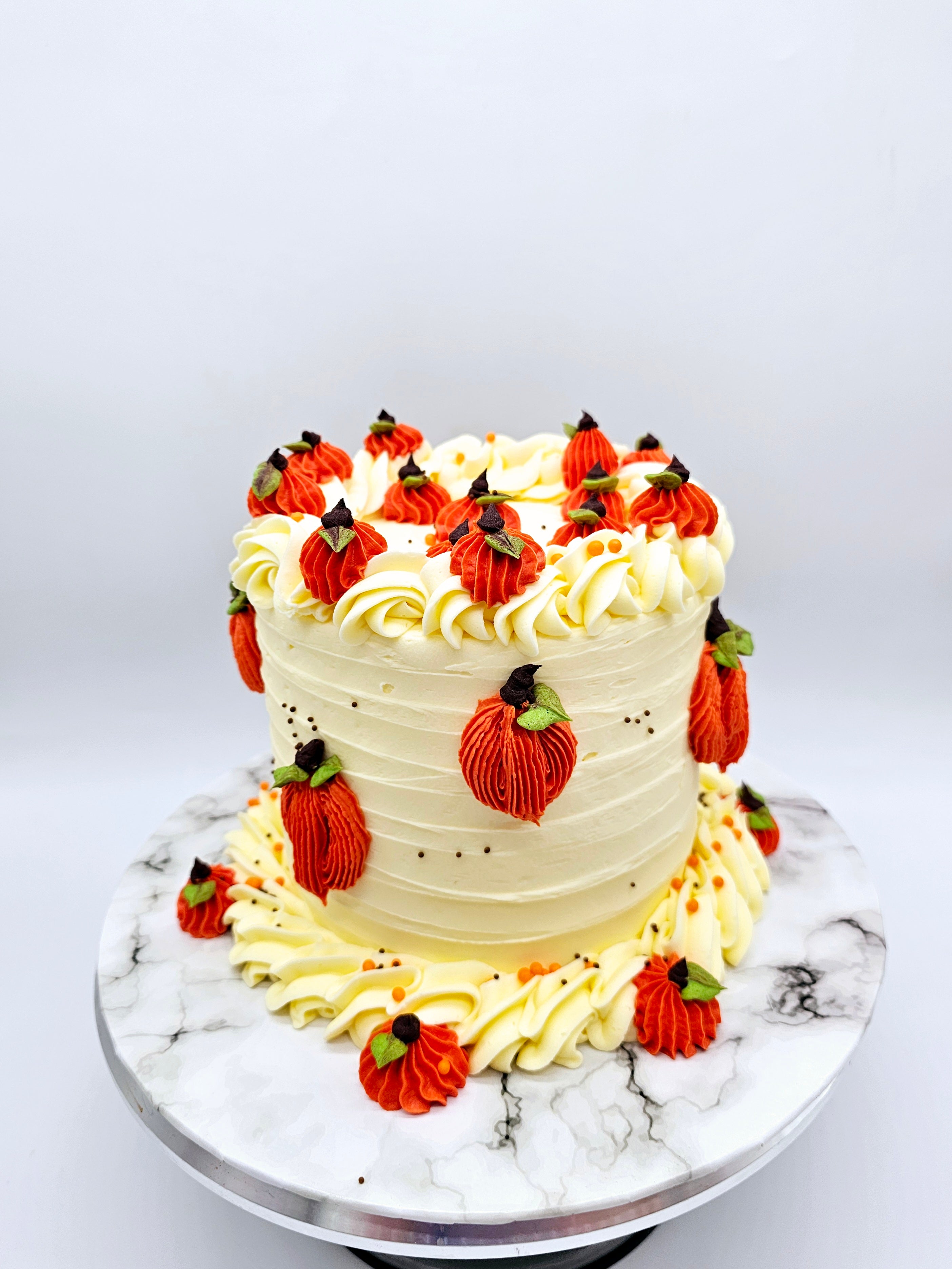 A pumpkin-themed autumn cake by Cake Secret, featuring piped orange pumpkins with green leaves and chocolate stems, surrounded by white buttercream frosting, displayed on a marble-effect cake stand.