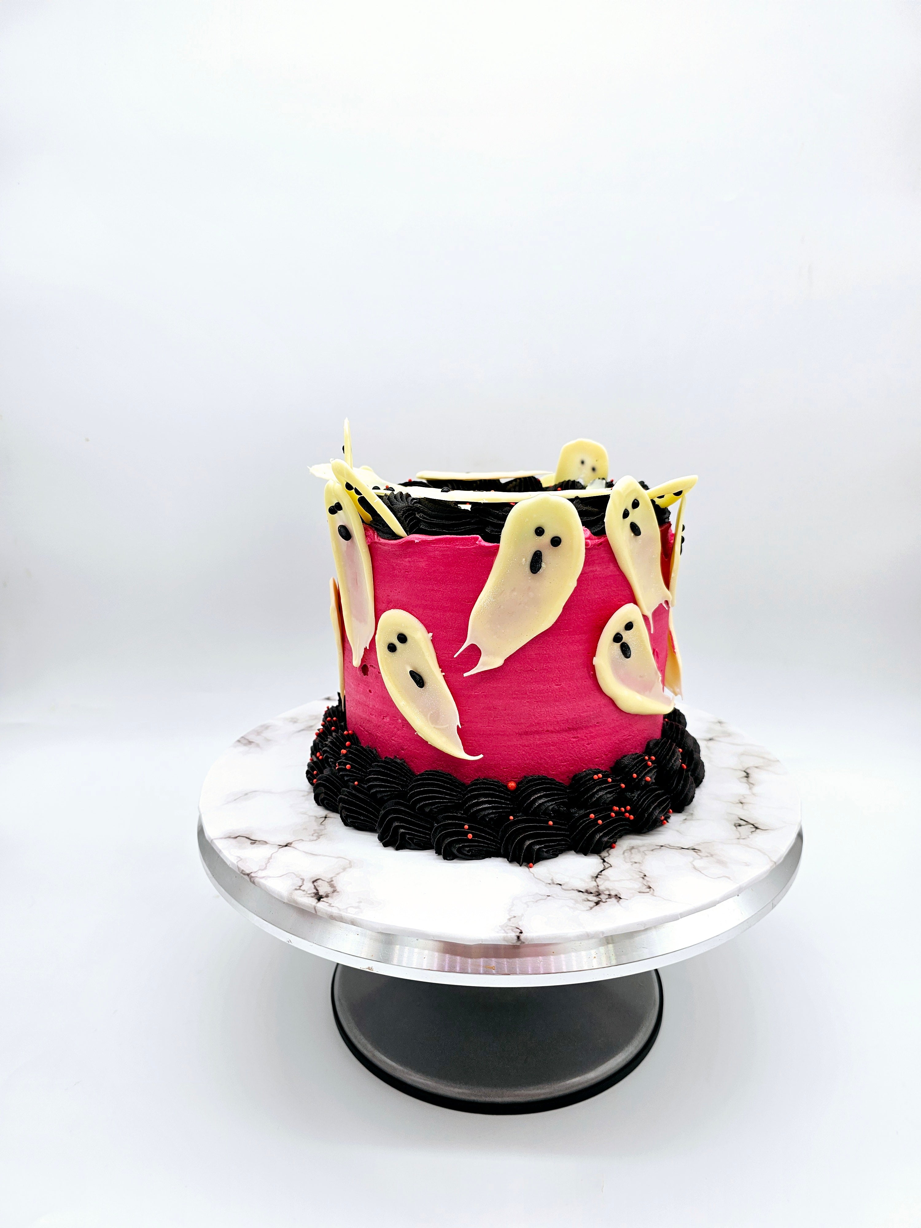 A Halloween-themed cake decorated with white chocolate ghost figures on a vibrant pink background, with black and red buttercream detailing, by Cake Secret.