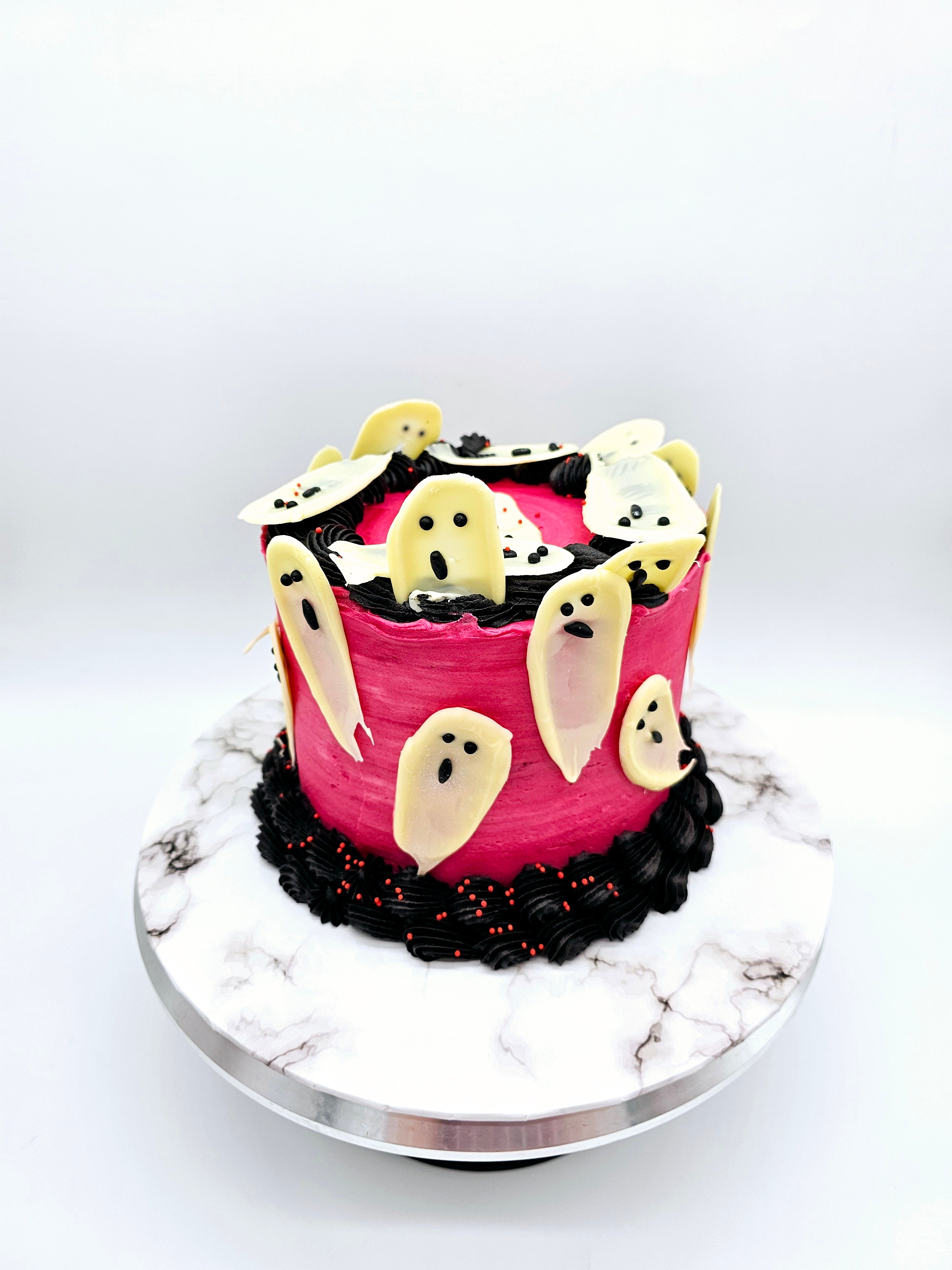 Top view of a Halloween-themed cake decorated with white chocolate ghost figures on a pink background, surrounded by black and red buttercream swirls, created by Cake Secret.