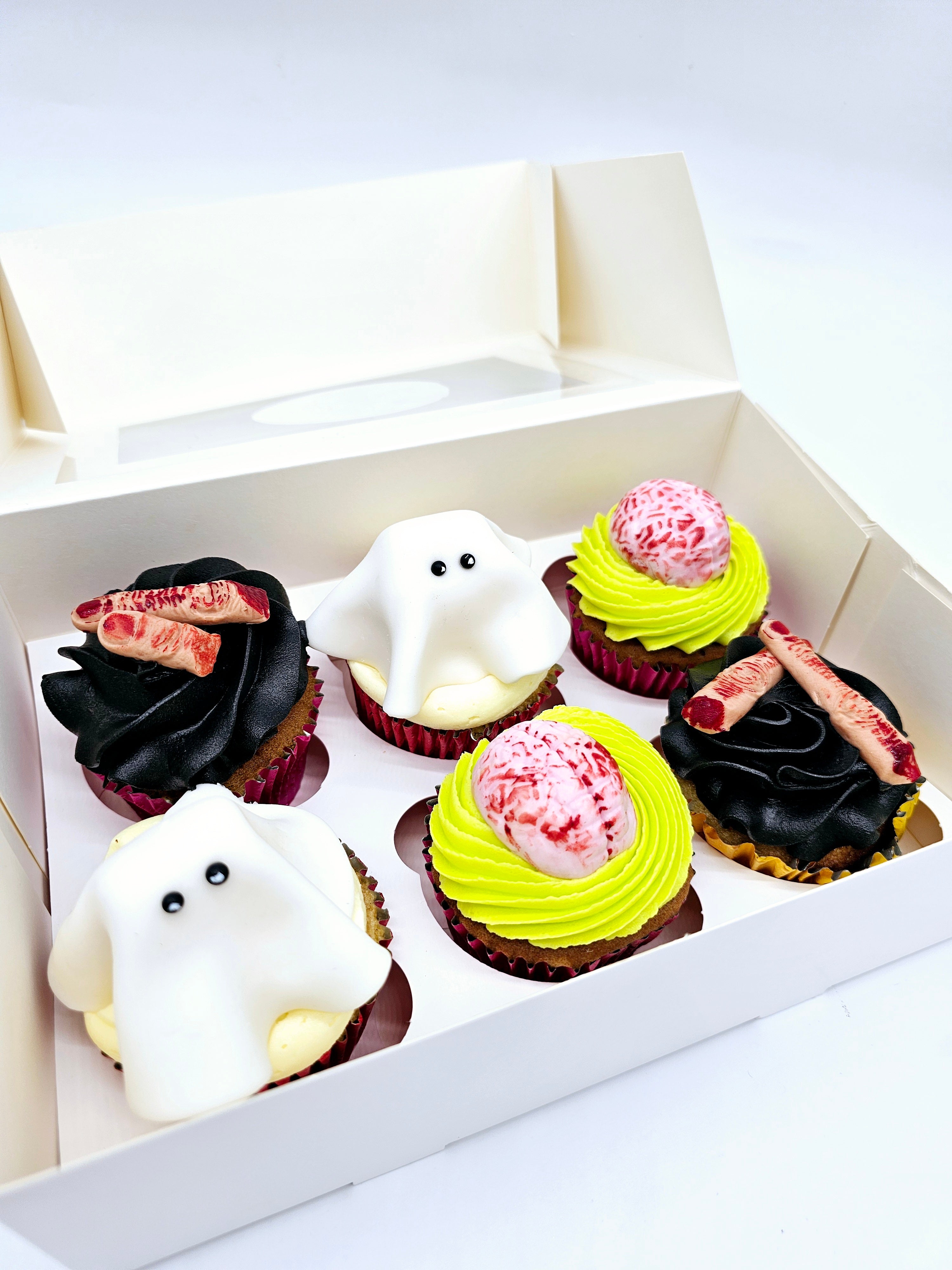 A box of six Halloween-themed cupcakes by Cake Secret, featuring ghost toppers, edible brains, and severed finger decorations on a mix of black and neon green buttercream, displayed in an open cupcake box.