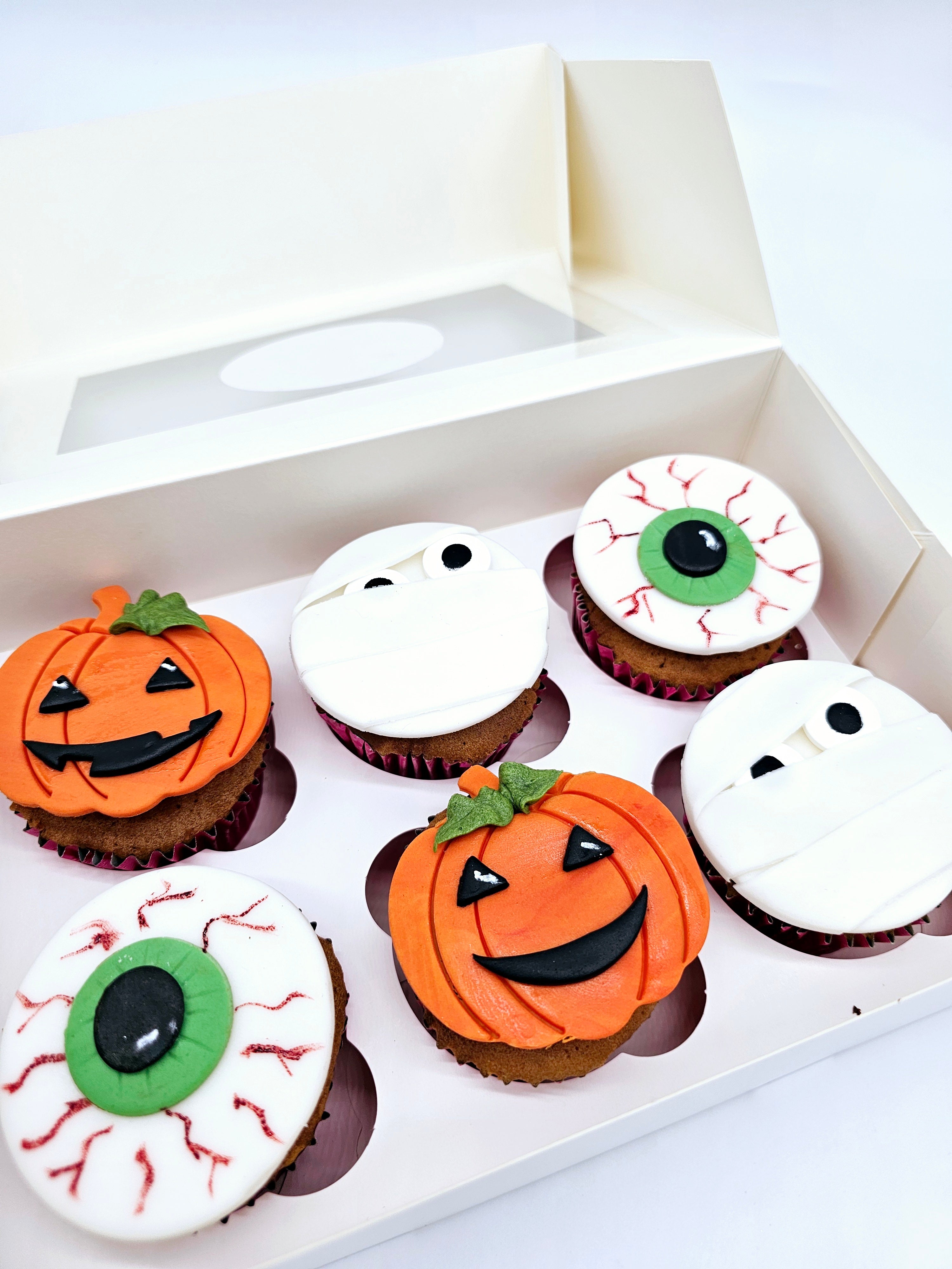 A box of six Halloween-themed cupcakes by Cake Secret, featuring fondant toppers shaped as pumpkins, mummies, and bloodshot eyeballs, presented in an open cupcake box