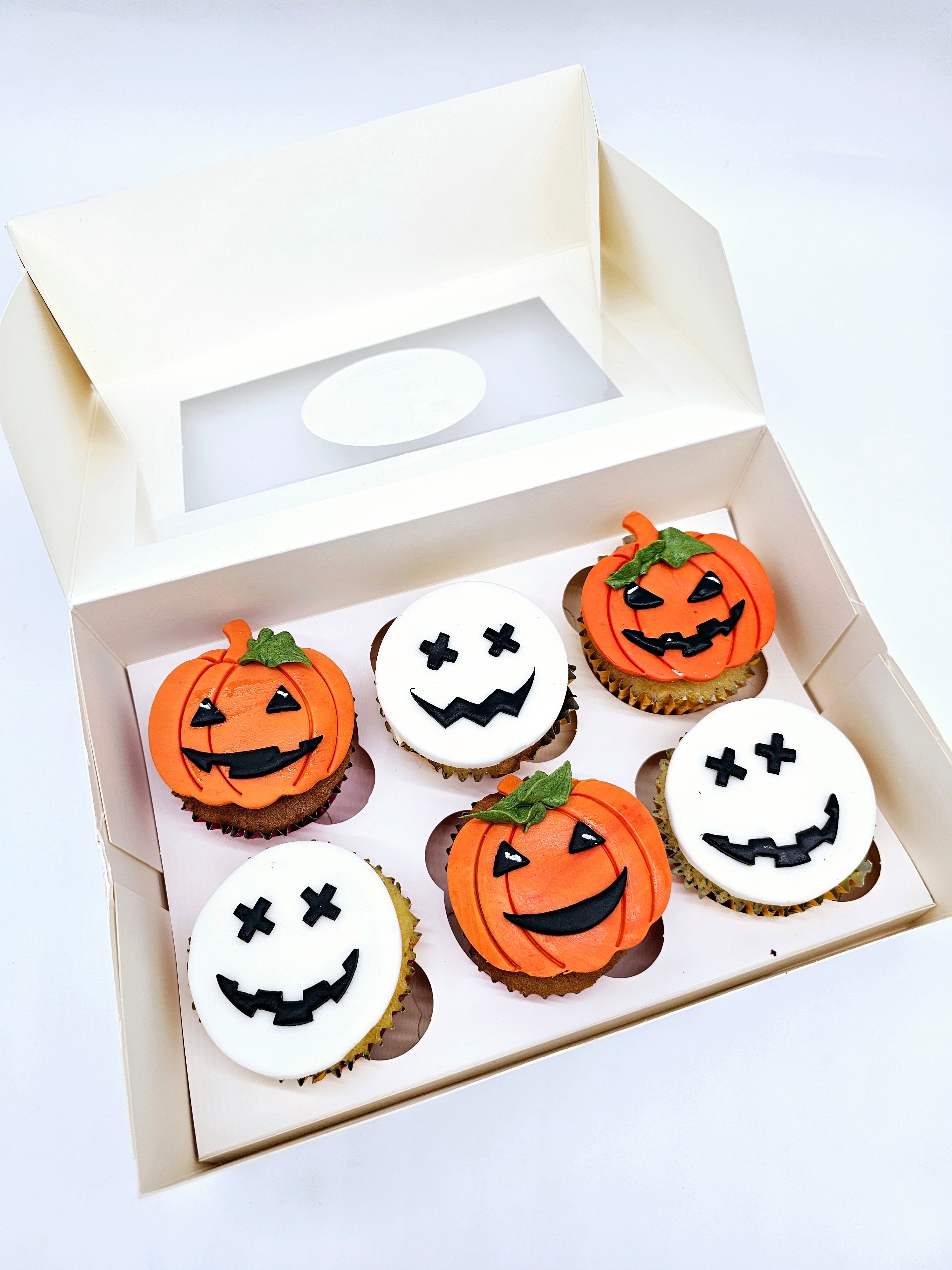 A box of six Halloween-themed cupcakes by Cake Secret, featuring a mix of orange pumpkin faces and white skull faces, each decorated with black icing expressions, displayed in an open cupcake box.