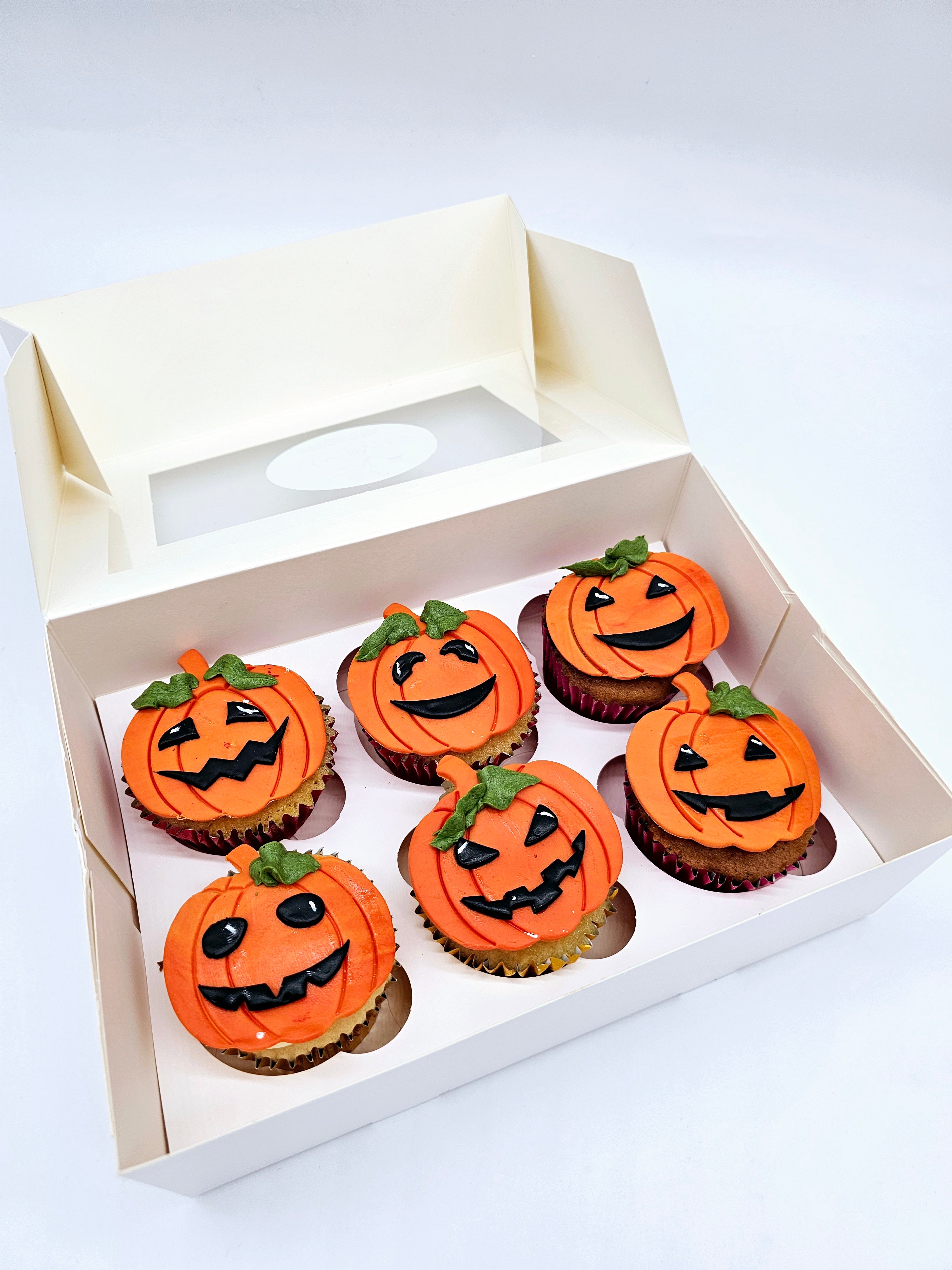 A box of six Halloween-themed cupcakes by Cake Secret, each decorated with orange pumpkin designs and various spooky expressions in black icing, displayed in an open cupcake box.