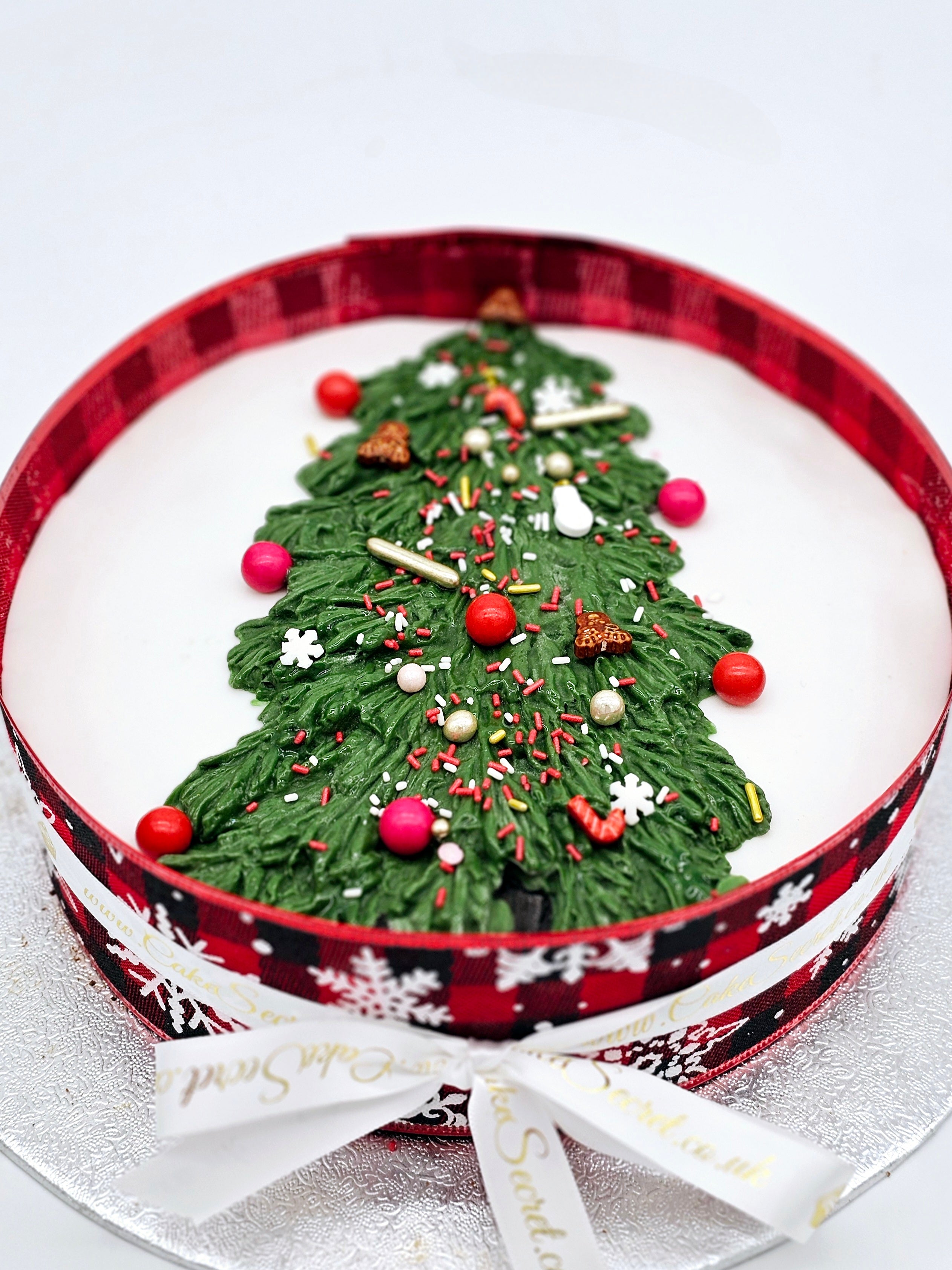 A close-up of a Christmas cake by Cake Secret, featuring a green piped Christmas tree design decorated with sprinkles, ornaments, and edible decorations, surrounded by a festive red plaid ribbon and snowflake-themed trim.
