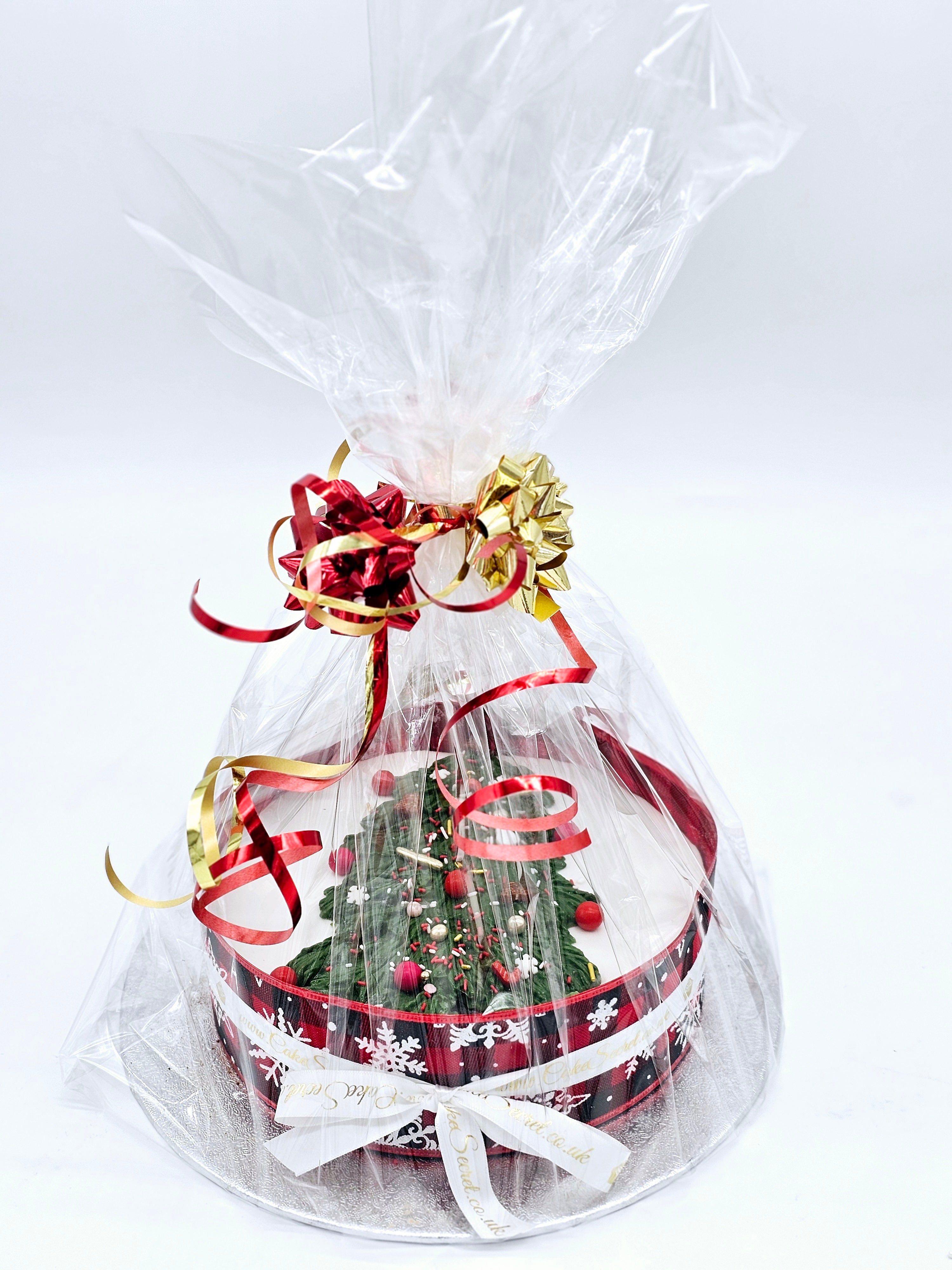 A Christmas cake by Cake Secret, packaged in a festive gift wrap with clear cellophane, decorated with red and gold ribbons, featuring a tree design and holiday decorations inside.