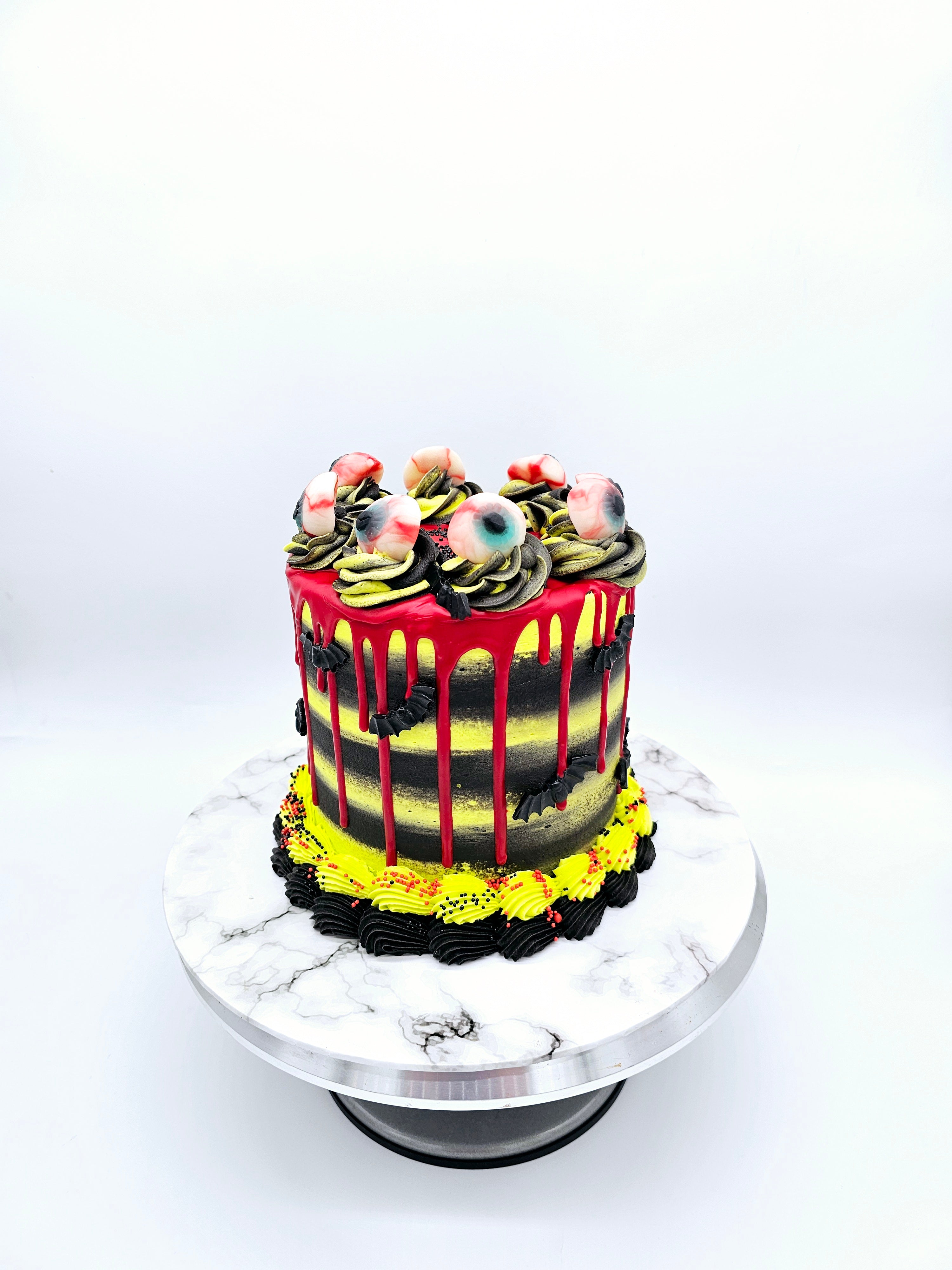A Halloween-themed cake by Cake Secret, featuring red drip icing, black and yellow buttercream layers, and decorated with edible eyeballs on top, displayed on a marble-effect cake stand.