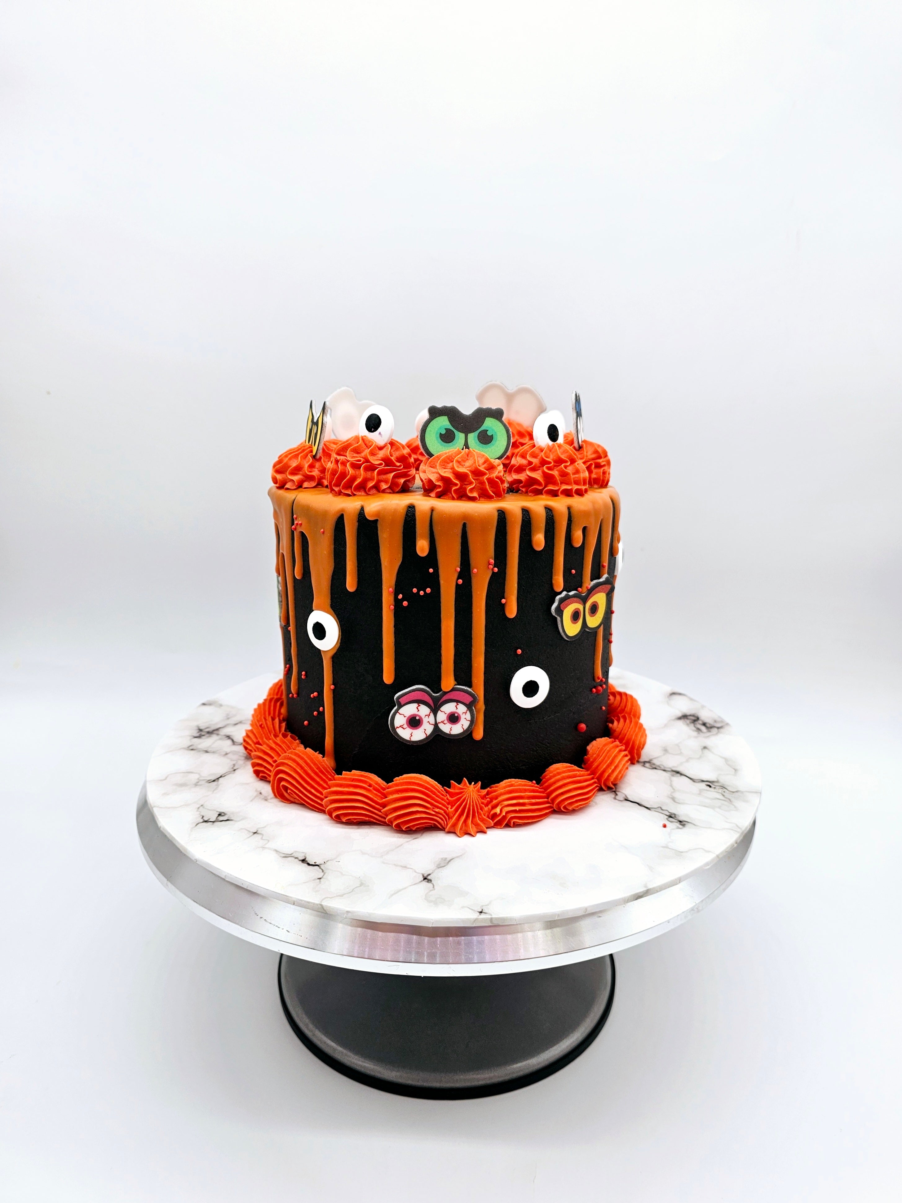 Front view of a Halloween-themed cake by Cake Secret, featuring orange drip icing over a black buttercream base, decorated with piped orange swirls, fondant eyes, ghost toppers, and spooky eyeball details, displayed on a marble-effect cake stand.