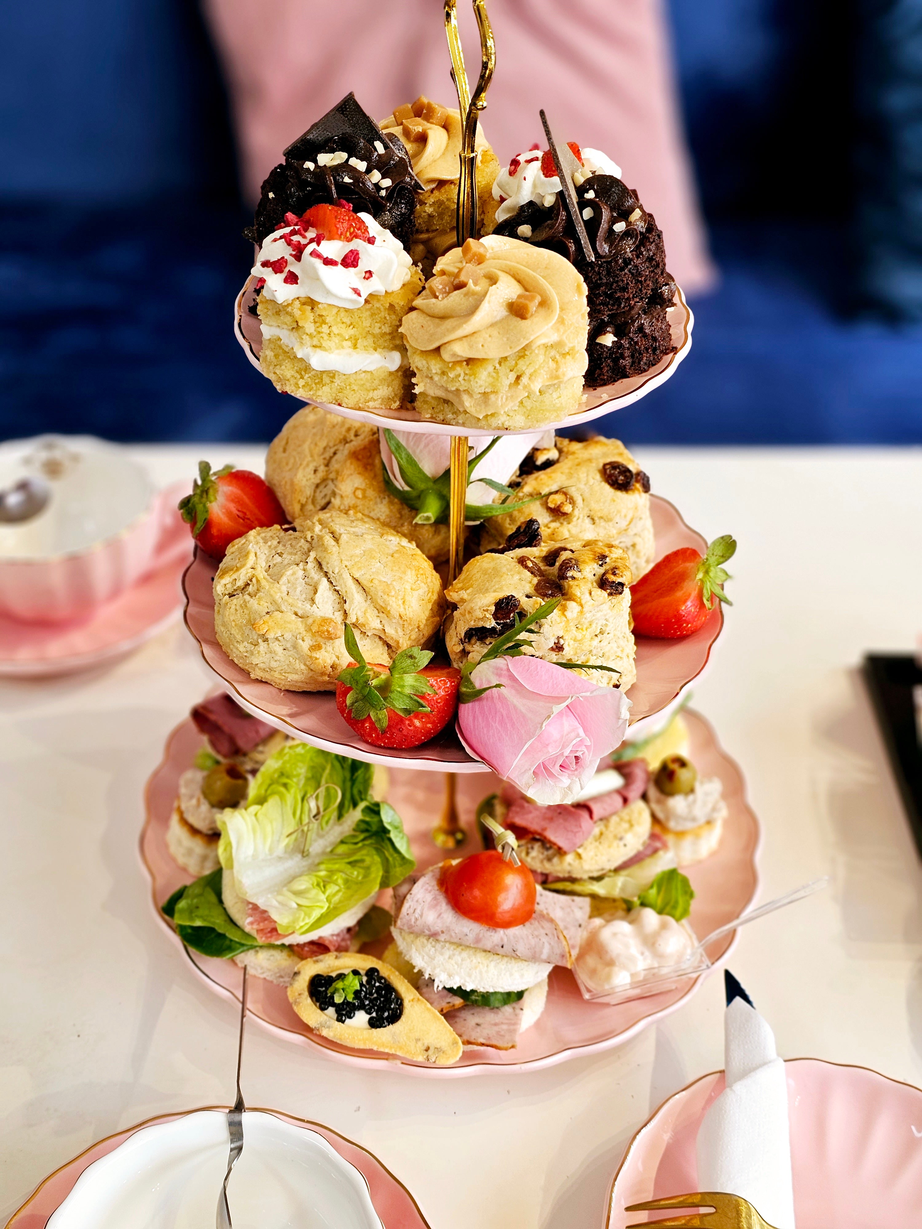 Front view of a three-tier cake stand from Cake Secret, showing scones, mini pastries, and fruit cakes on display for afternoon tea, with the table and surrounding décor visible.