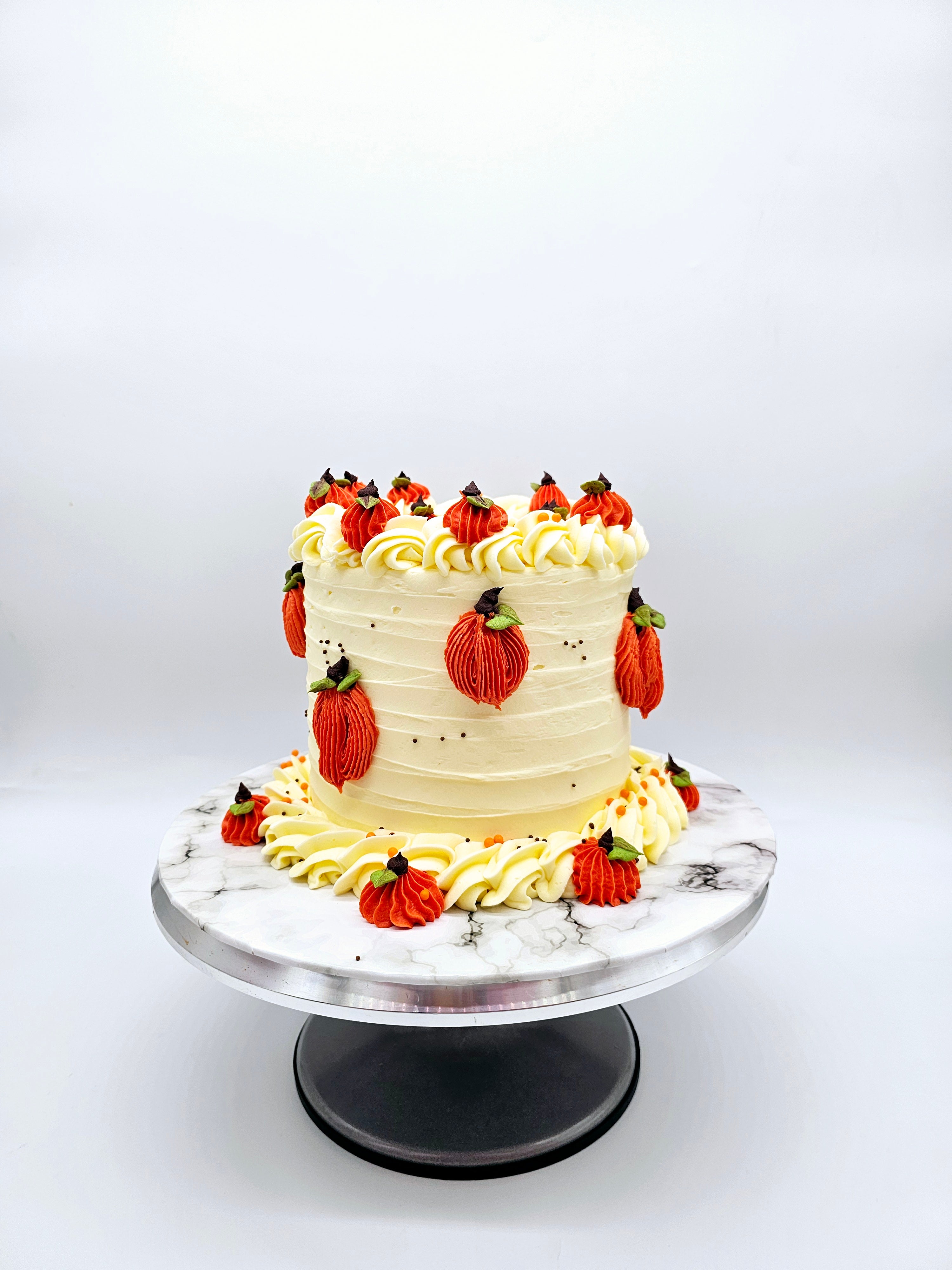 Front view of a pumpkin-themed autumn cake by Cake Secret, featuring piped orange pumpkins with green leaves and chocolate stems, topped with white buttercream swirls, displayed on a marble-effect cake stand.