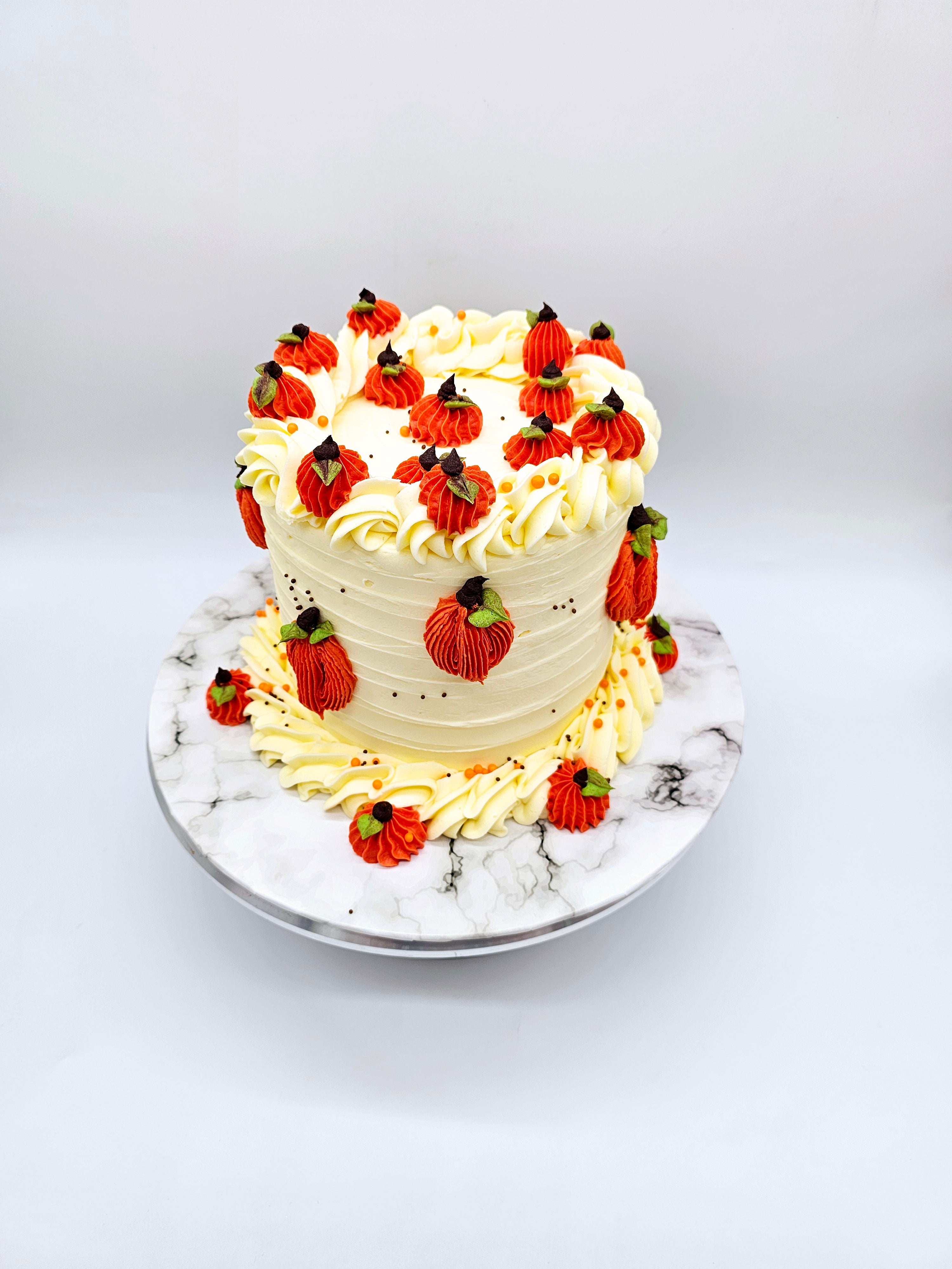 Top view of a pumpkin-themed autumn cake by Cake Secret, featuring piped orange pumpkins with green leaves and chocolate stems, surrounded by white buttercream swirls, displayed on a marble-effect cake stand.