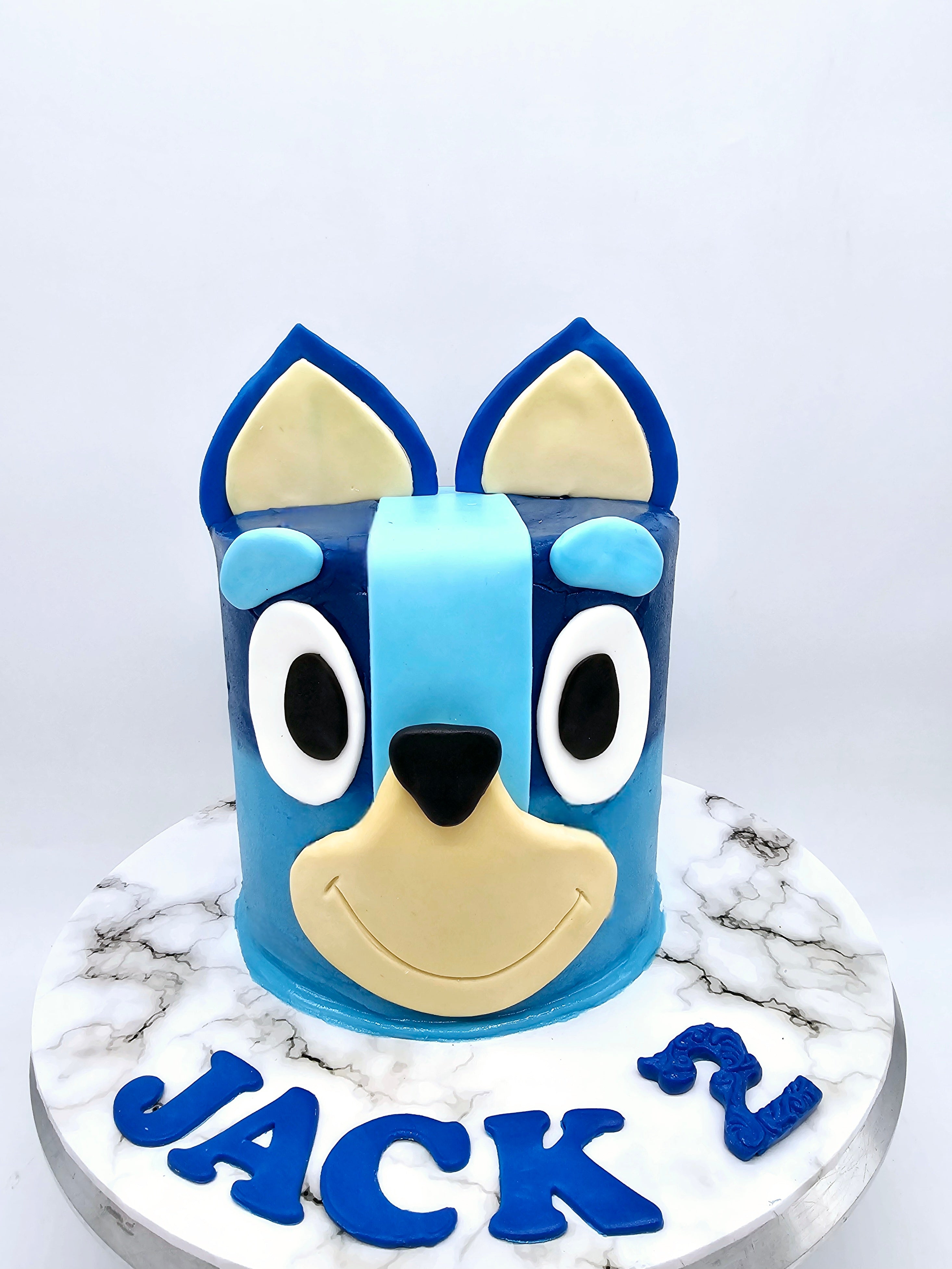 Front view of a Bluey-themed second birthday cake made by Cake Secret, featuring the 3D face of Bluey in blue and beige fondant, with the name Jack and the number two in blue fondant, displayed on a marble-effect cake stand.