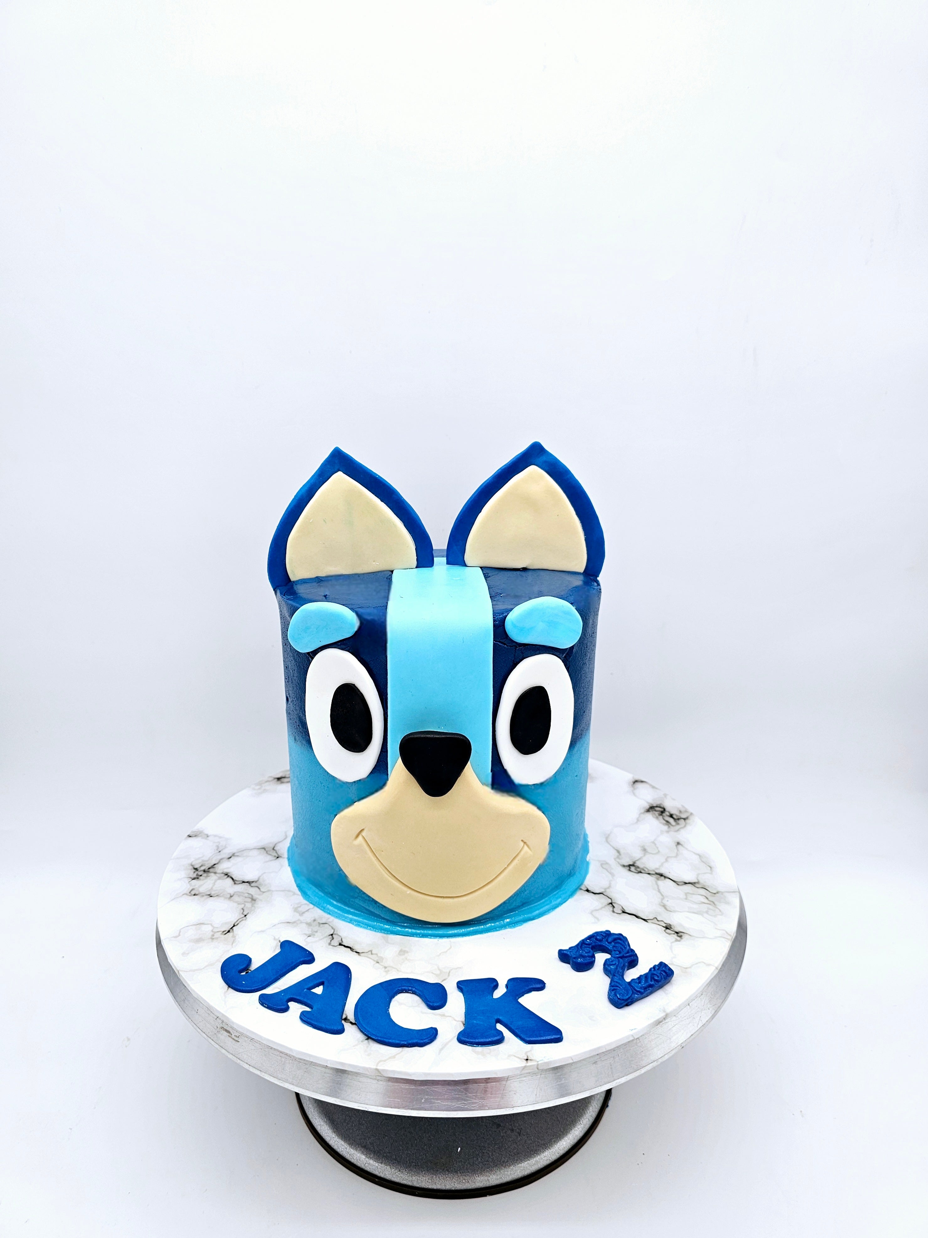 Full front view of a Bluey-themed second birthday cake made by Cake Secret, featuring the 3D face of Bluey in blue and beige fondant, with the name Jack and the number two in blue fondant, displayed on a marble-effect cake stand.