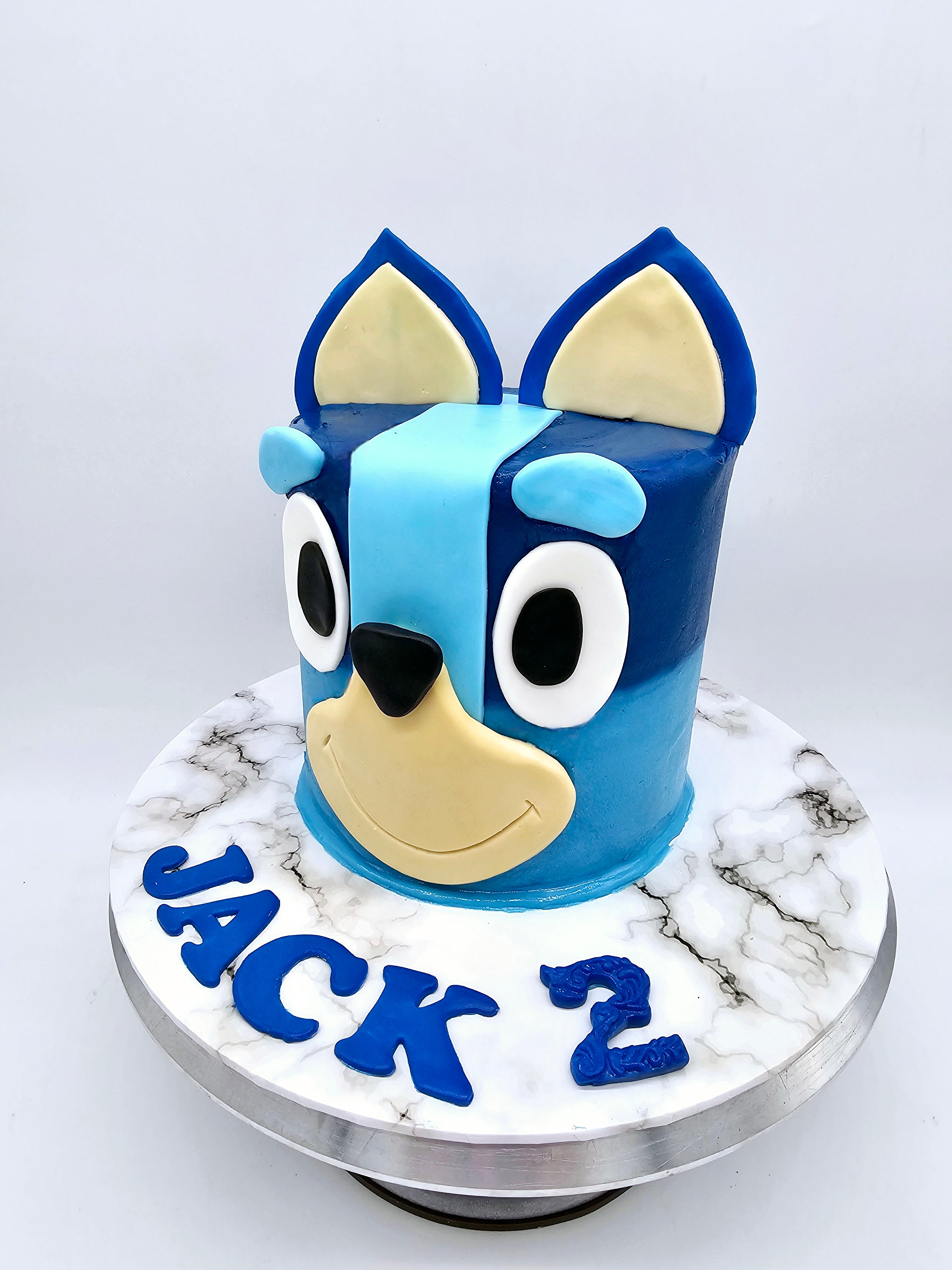 A Bluey-themed second birthday cake made by Cake Secret, featuring the 3D face of Bluey, with blue and beige fondant details, and the name Jack and the number two in blue fondant, displayed on a marble-effect cake stand.