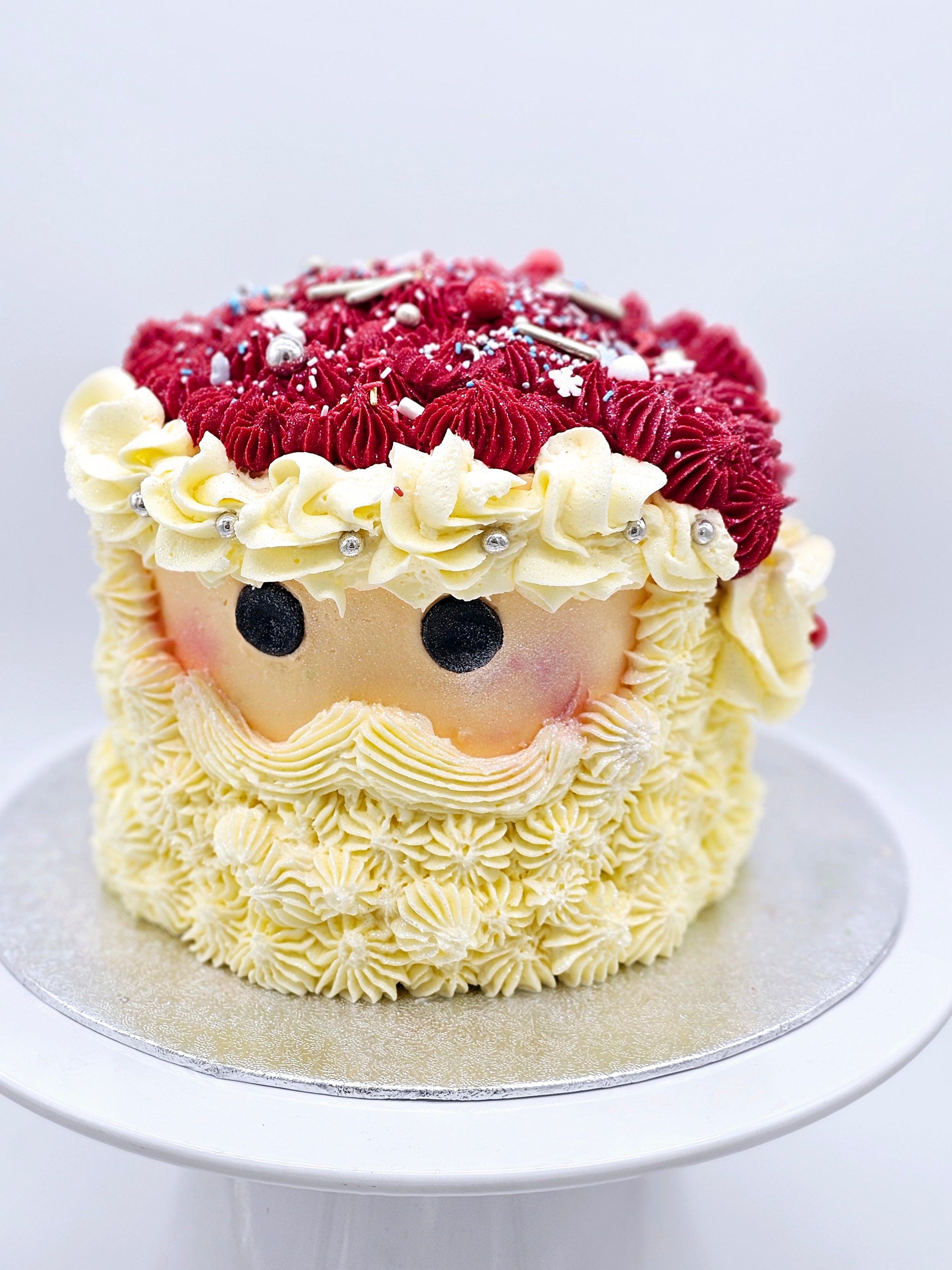 A Santa-themed Christmas cake by Cake Secret, featuring a detailed buttercream design with Santa’s face, red piped icing for the hat, and white piped icing for the beard, decorated with sprinkles and edible silver accents, displayed on a white cake stand.