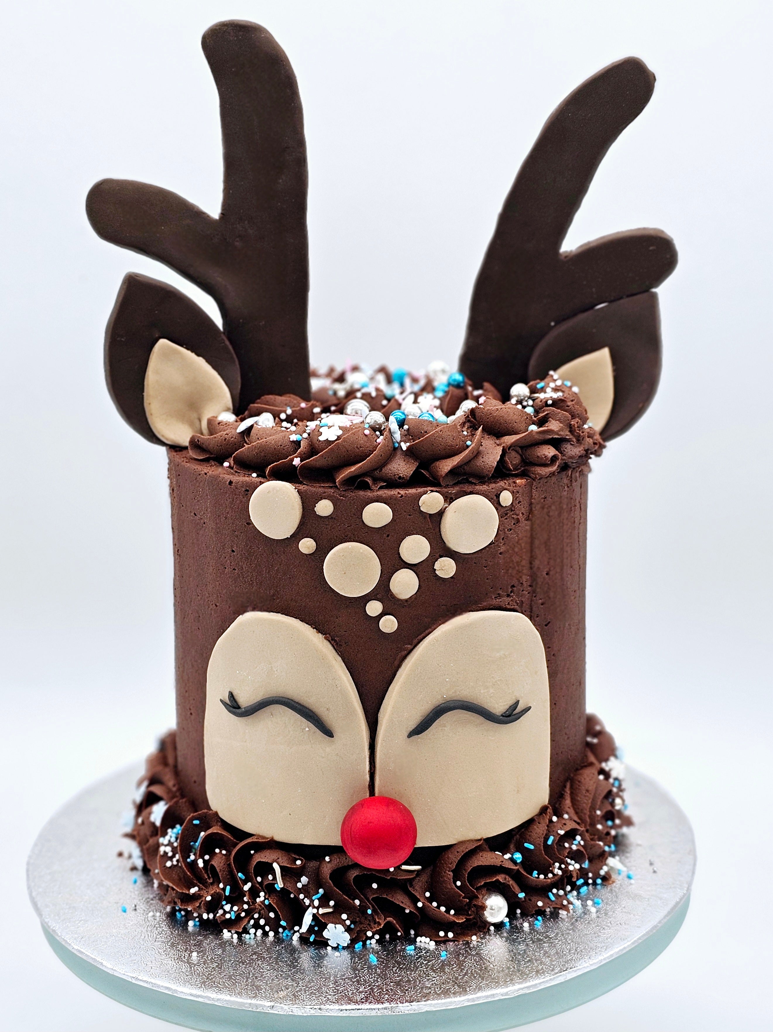A reindeer-themed Christmas cake by Cake Secret, featuring a chocolate buttercream design with fondant reindeer antlers, ears, and a red nose, decorated with sprinkles and edible silver accents, displayed on a silver cake board.