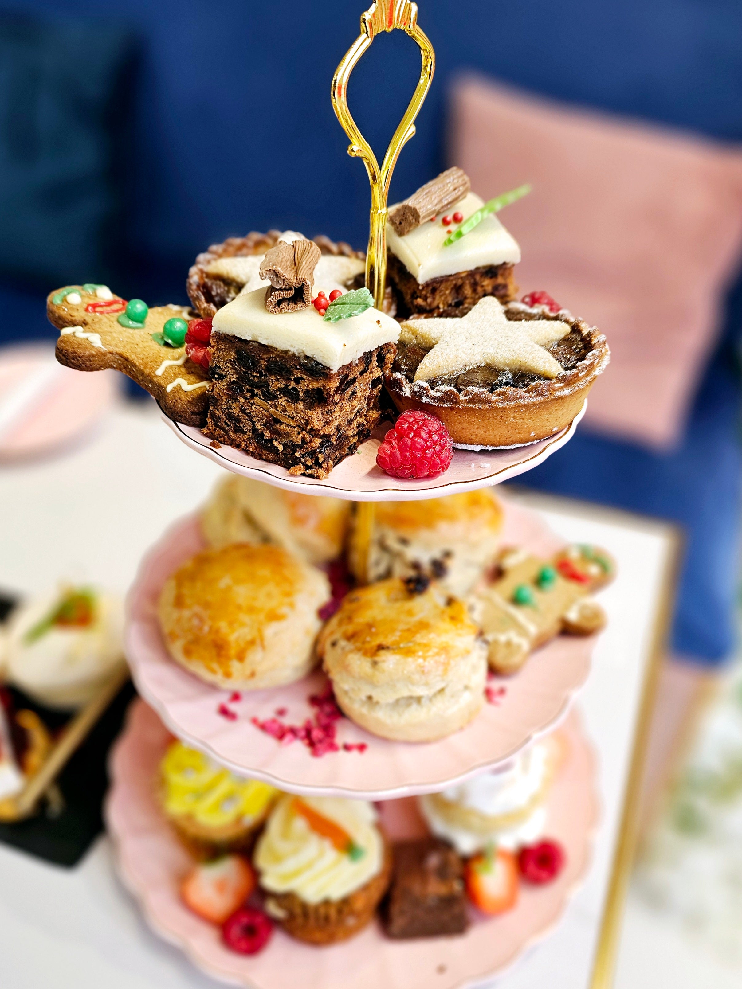 Front view of a three-tier cake stand from Cake Secret showcasing Christmas afternoon tea items, including festive fruit cakes, mince pies, scones, and gingerbread cookies, arranged with fresh raspberries and holiday decorations.