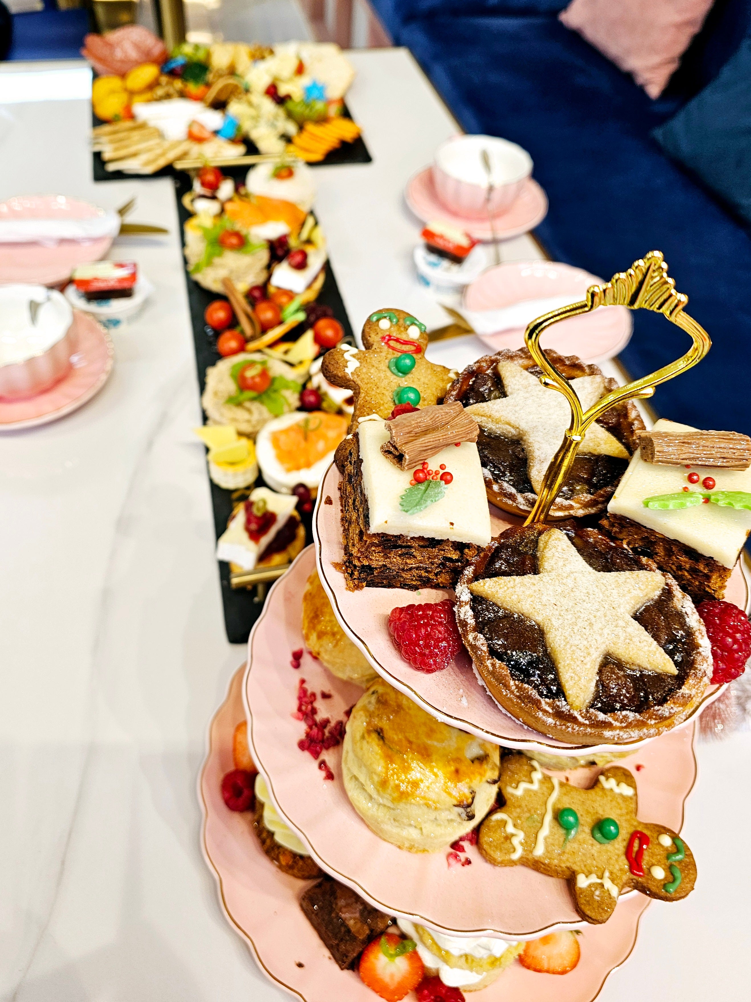 Long view of a three-tier cake stand from Cake Secret showcasing Christmas afternoon tea items, including festive fruit cakes, mince pies, scones, and gingerbread cookies, arranged with fresh raspberries and holiday decorations.