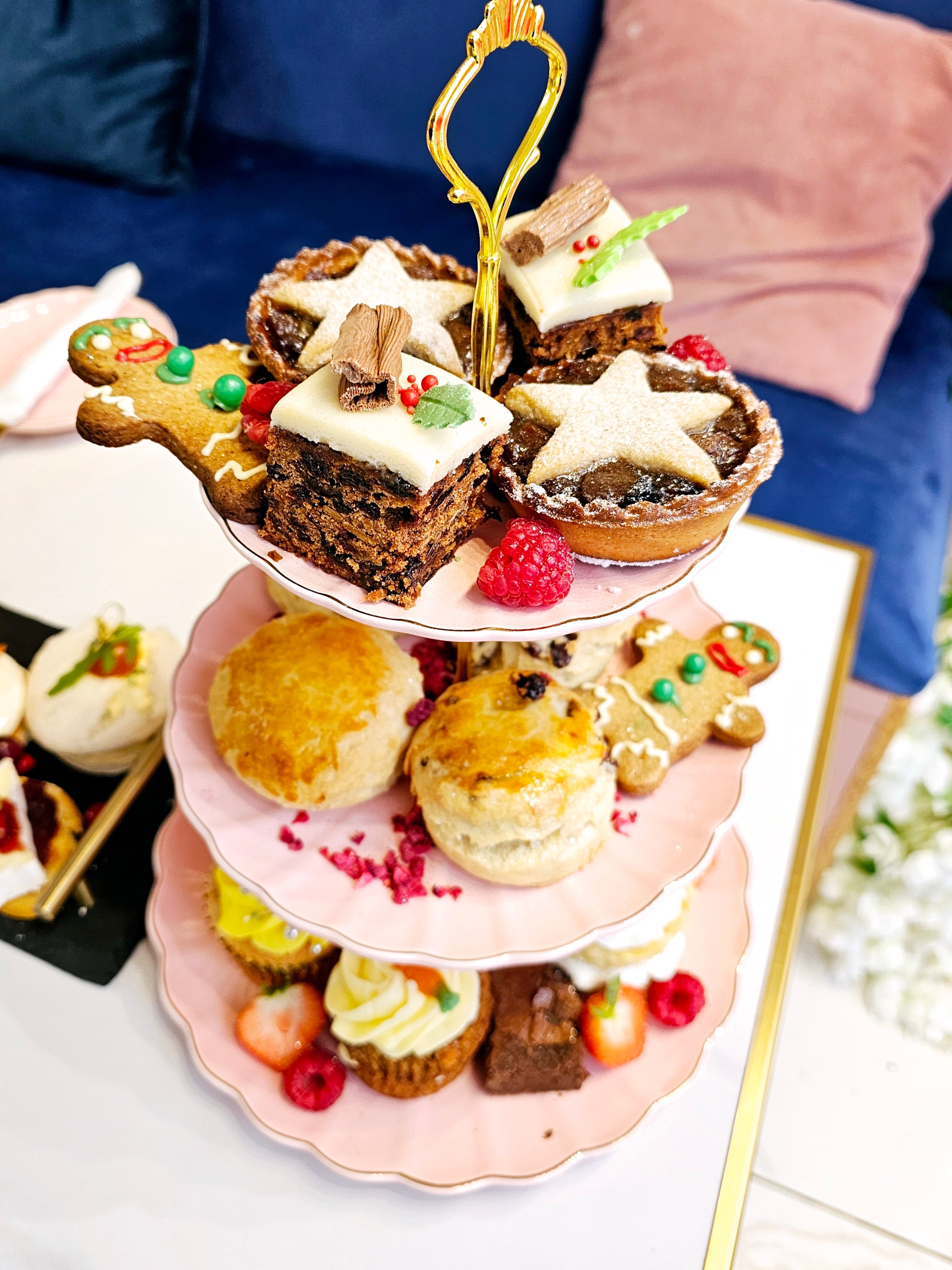 Over view of a three-tier cake stand from Cake Secret showcasing Christmas afternoon tea items, including festive fruit cakes, mince pies, scones, and gingerbread cookies, arranged with fresh raspberries and holiday decorations.