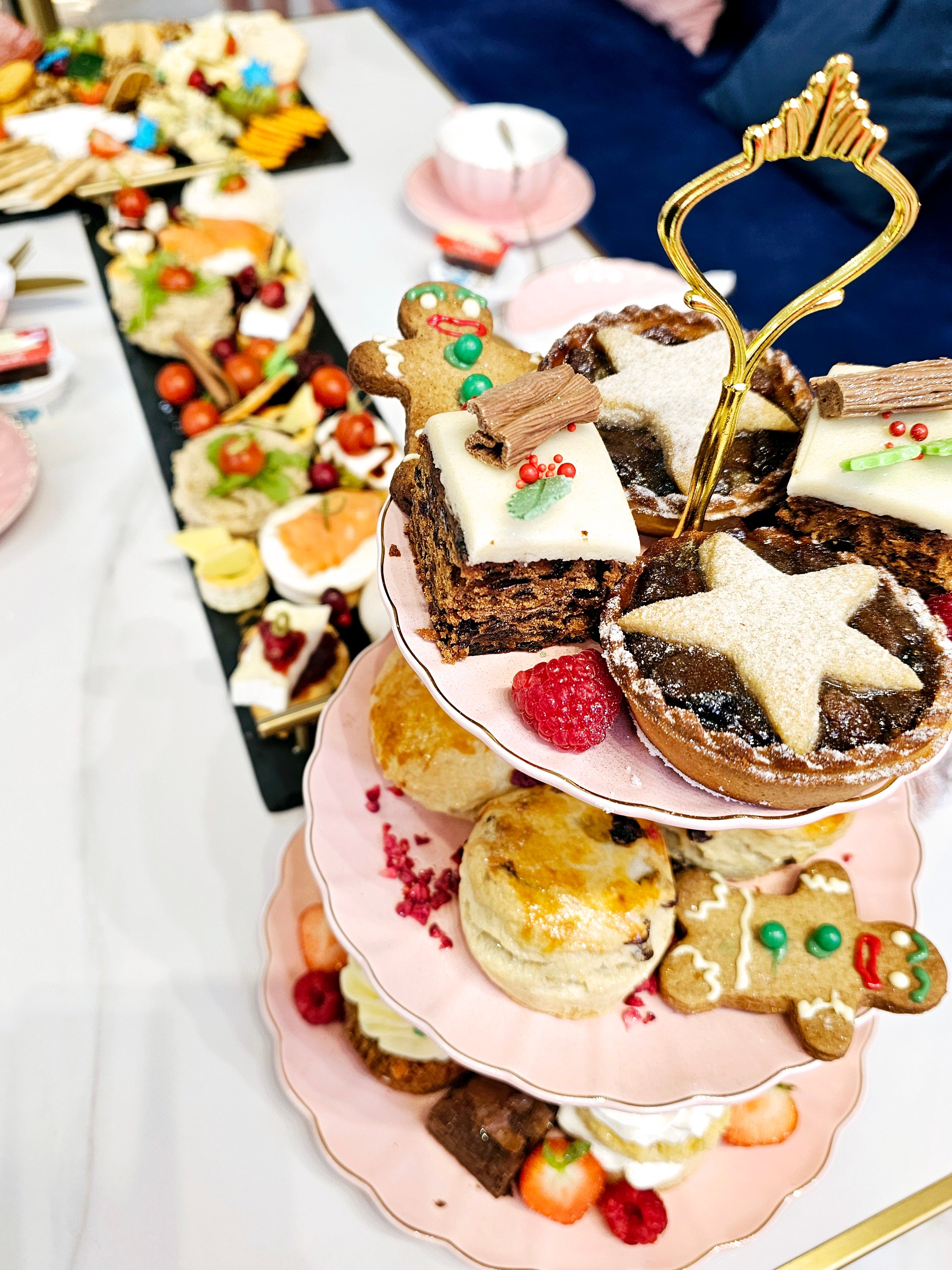 Wide view of a three-tier cake stand from Cake Secret showcasing Christmas afternoon tea items, including festive fruit cakes, mince pies, scones, and gingerbread cookies, arranged with fresh raspberries and holiday decorations.