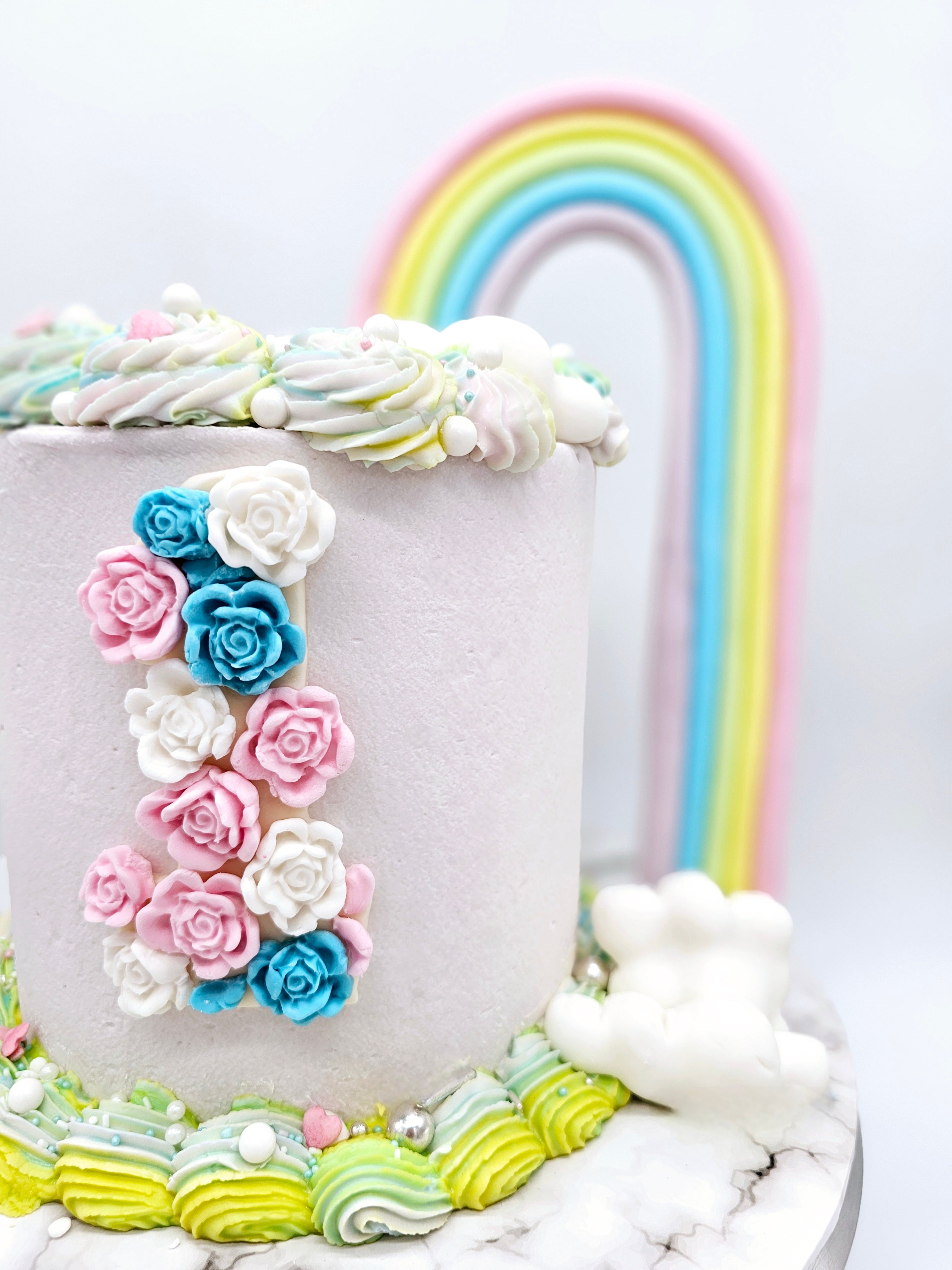 Close-up of a pastel-coloured first birthday cake made by Cake Secret, featuring a fondant rainbow arch, floral decorations forming the number one, and piped icing details on a marble-effect cake stand.