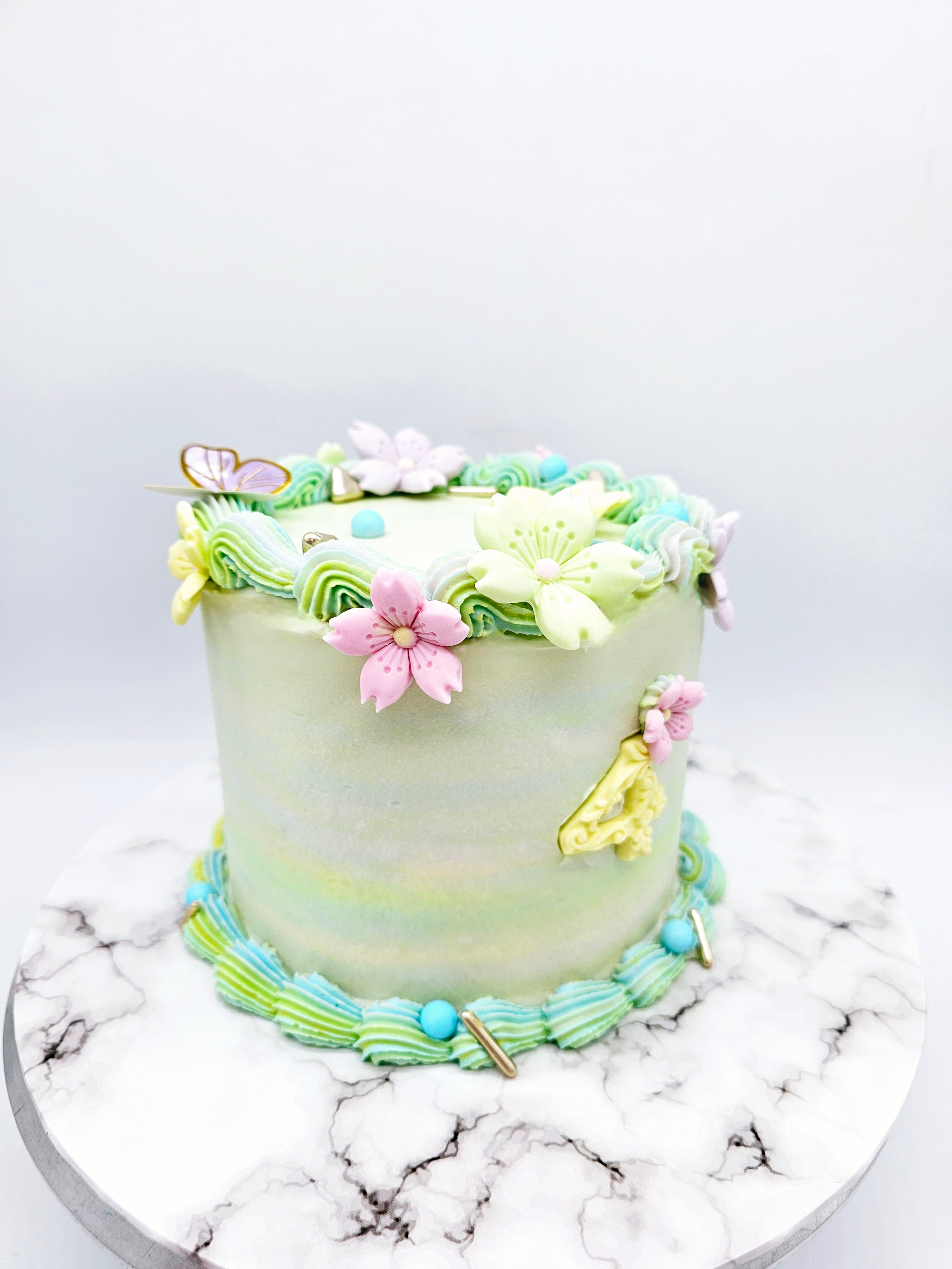 Close side view of a pastel-coloured 4th birthday cake made by Cake Secret, decorated with piped floral icing details, a gold number four, and pastel buttercream swirls, displayed on a marble-effect cake stand.