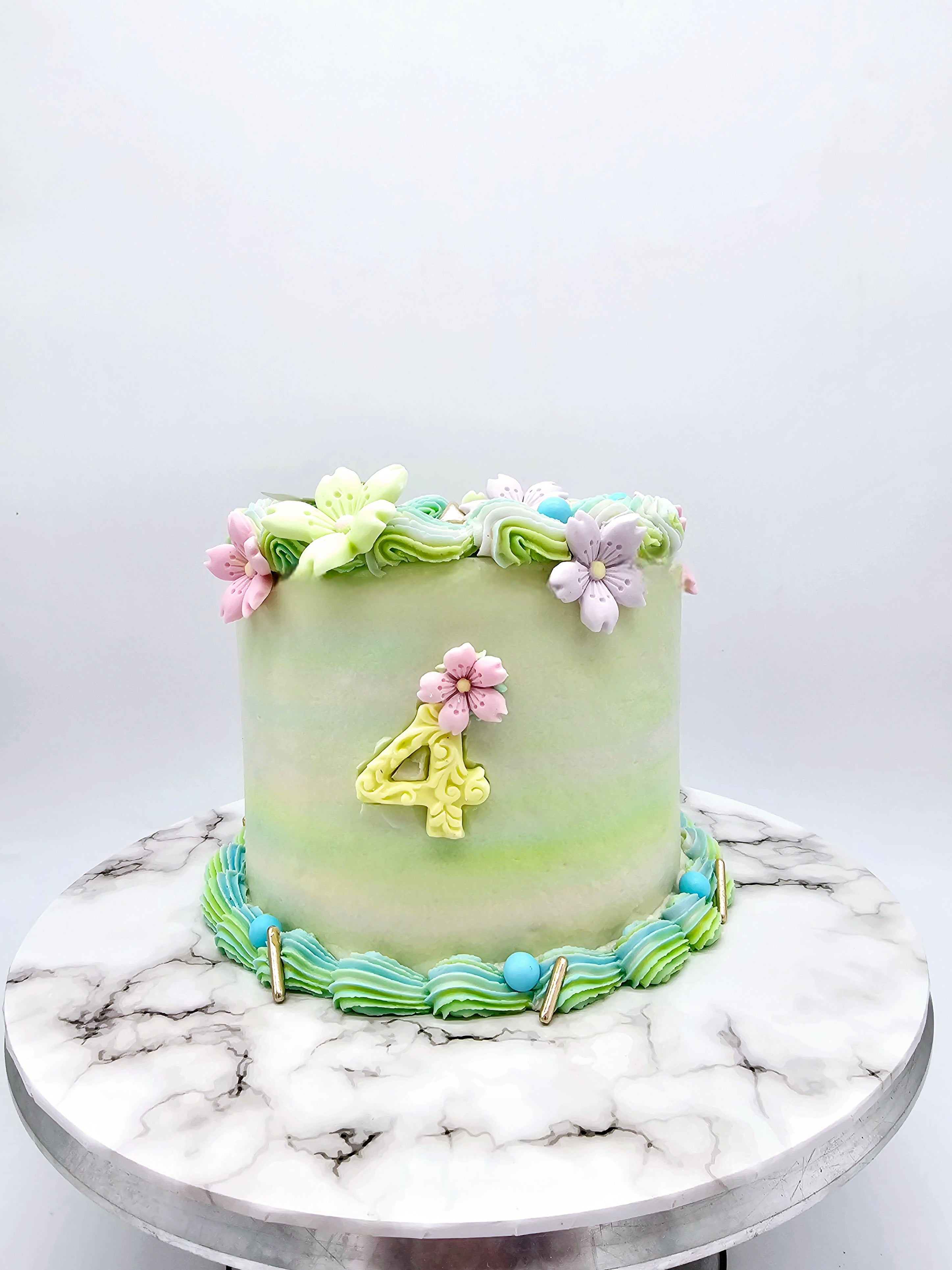 Front view of a pastel-coloured 4th birthday cake made by Cake Secret, featuring piped floral icing, a gold number four, and buttercream swirls around the base, displayed on a marble-effect cake stand.