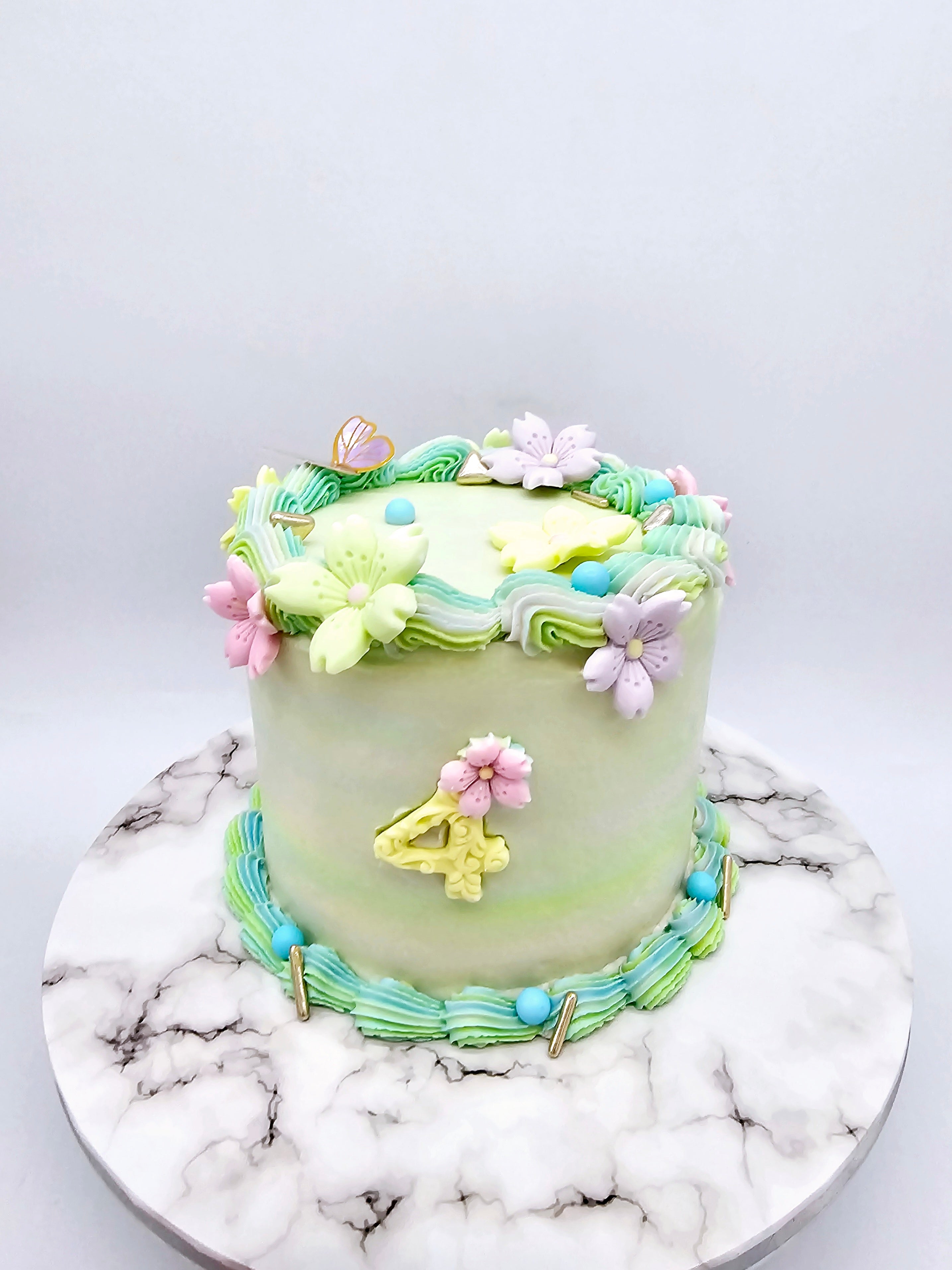 Side view of a pastel-coloured 4th birthday cake made by Cake Secret, featuring piped floral icing details, a gold number four, and pastel buttercream accents around the base, displayed on a marble-effect cake stand.