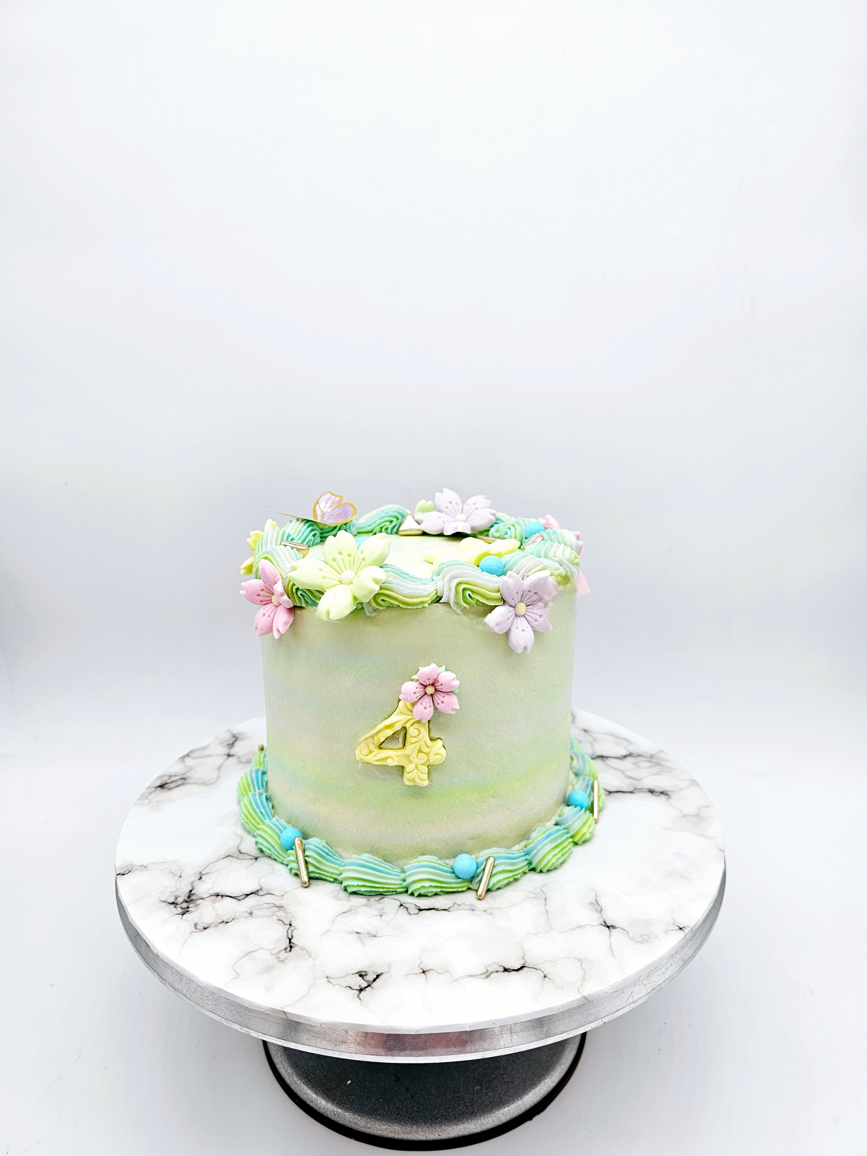 A pastel-coloured 4th birthday cake made by Cake Secret, decorated with piped floral icing details, a gold number four, and pastel buttercream accents, displayed on a marble-effect cake stand.