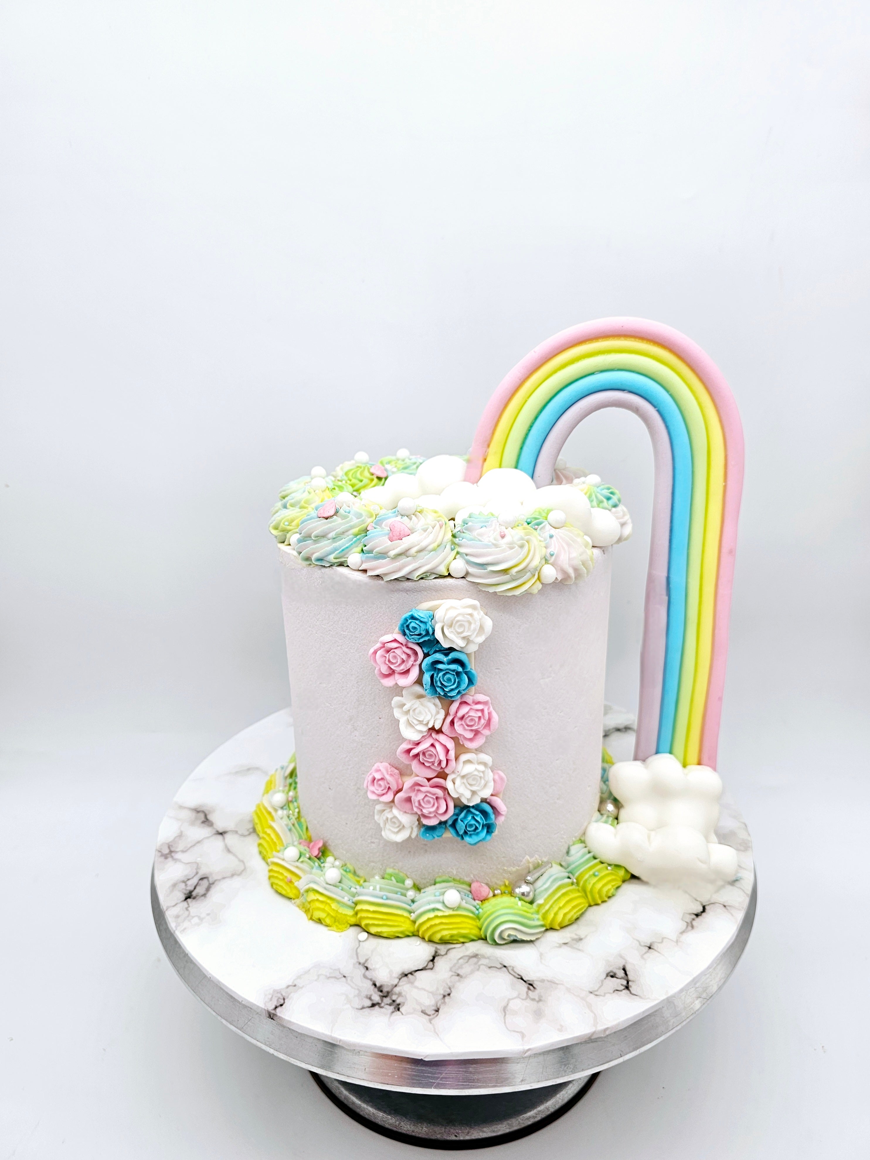 A pastel-coloured first birthday cake made by Cake Secret, featuring a fondant rainbow arch, floral decorations forming the number one, and piped icing details along the base, displayed on a marble-effect cake stand.