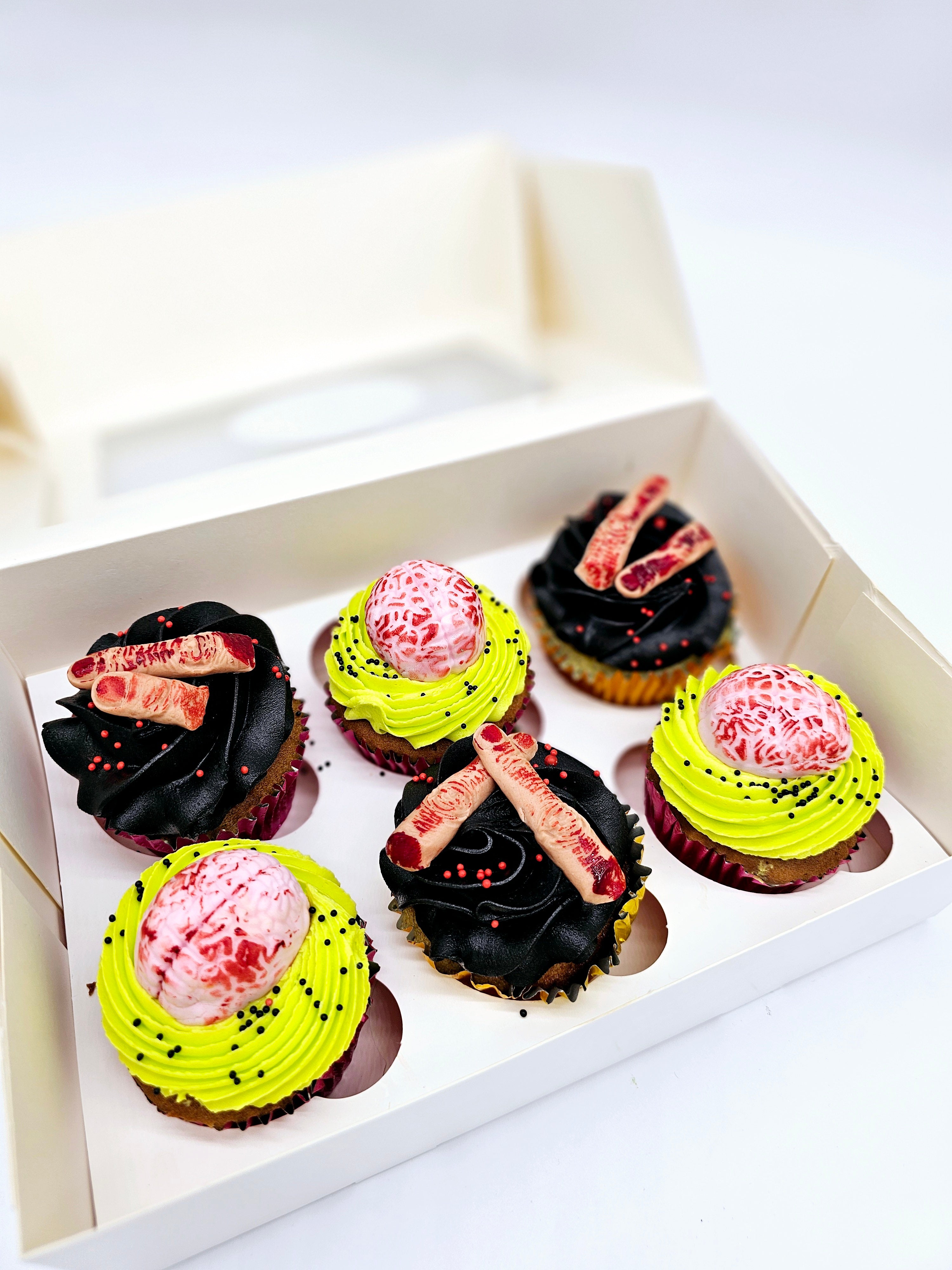 Six Halloween cupcakes by Cake Secret, decorated with edible brains on neon green frosting and severed fingers on black icing, arranged in a white box with spooky sprinkles.