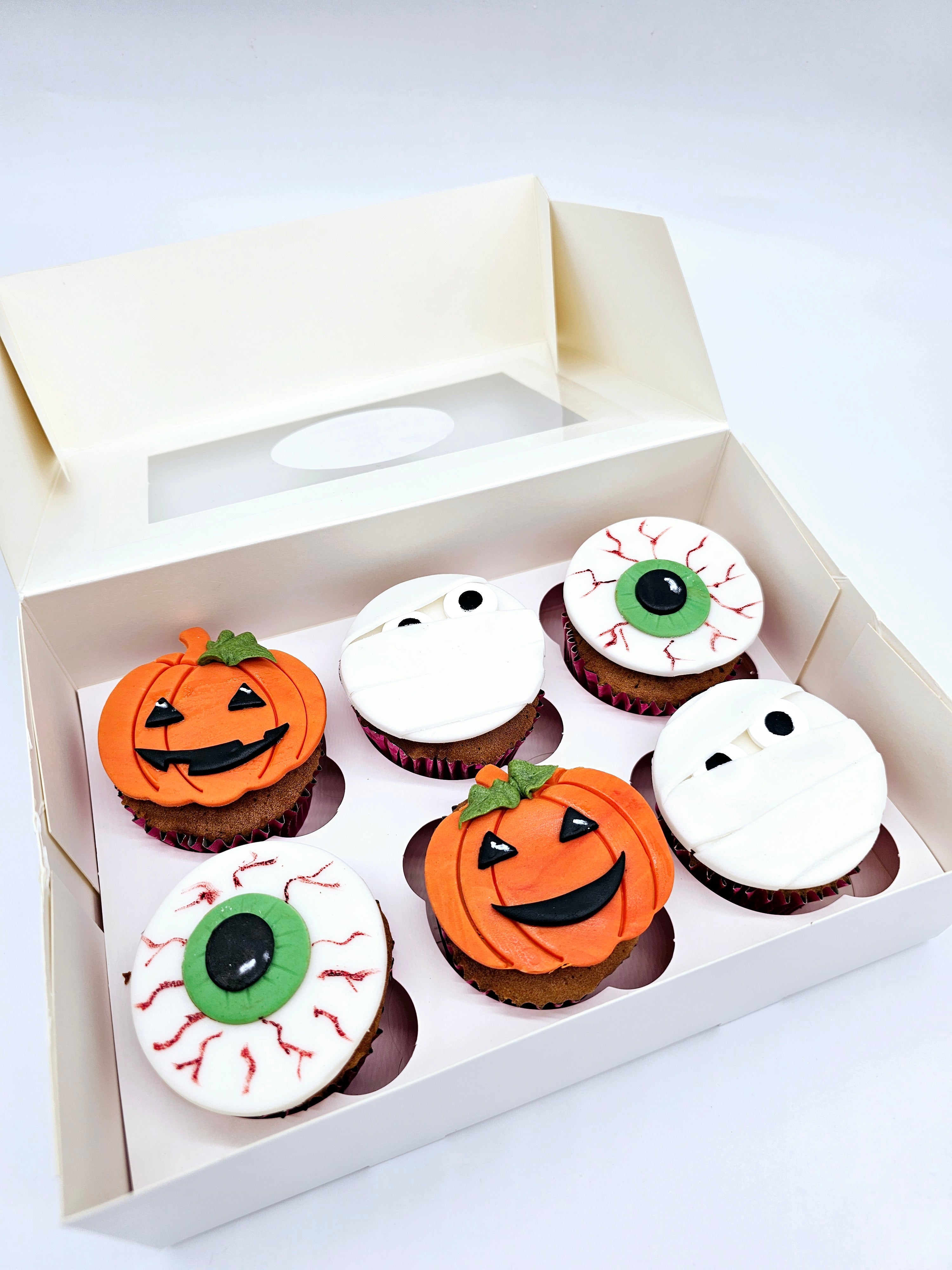 Six Halloween-themed cupcakes in a white box, decorated with fondant designs of pumpkins, mummies, and eyeballs, perfect for the spooky season by Cake Secret.