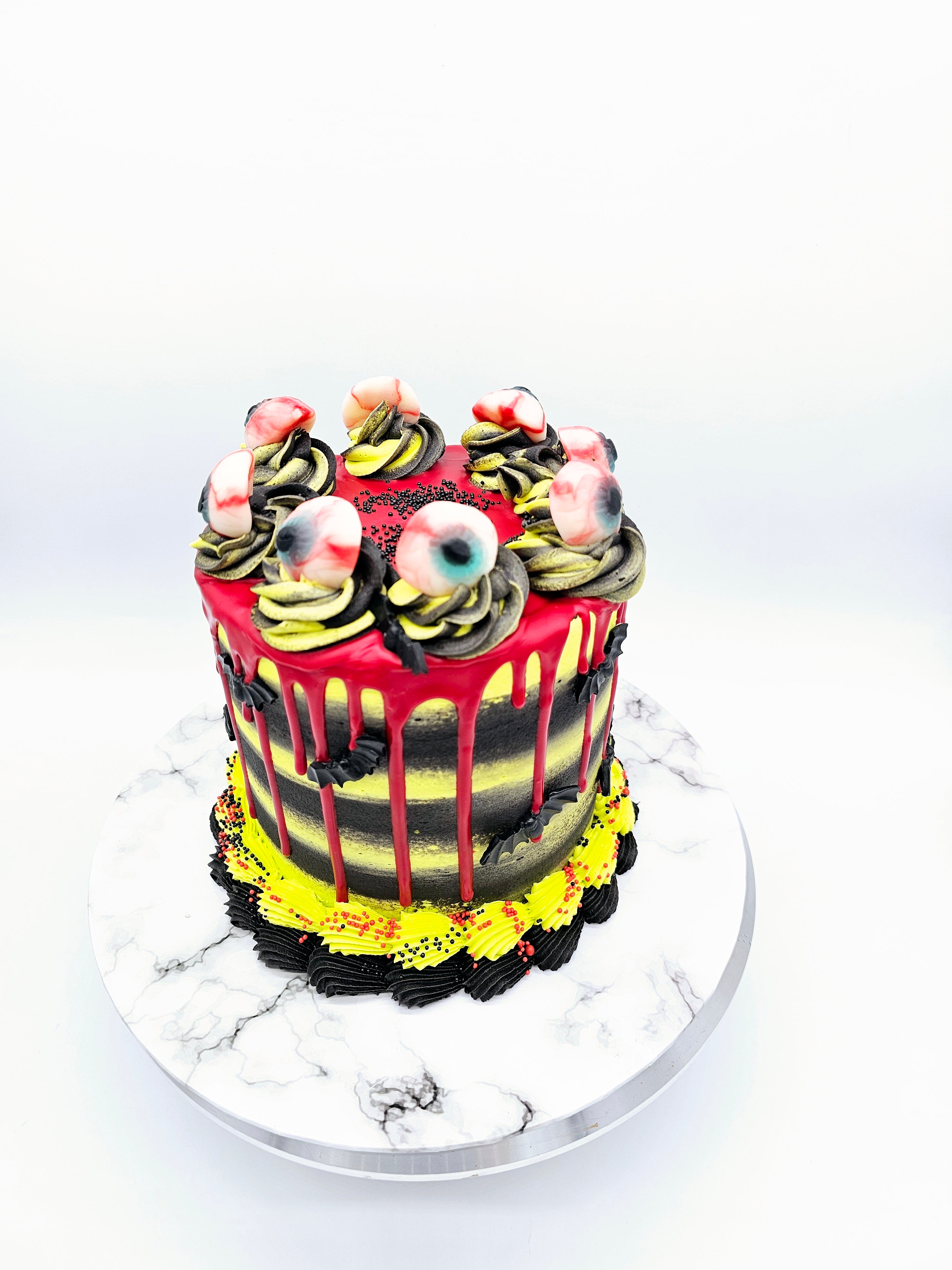 Top view of a Halloween-themed cake by Cake Secret, featuring red drip icing, black and yellow buttercream layers, and decorated with edible eyeballs on top, displayed on a marble-effect cake stand.