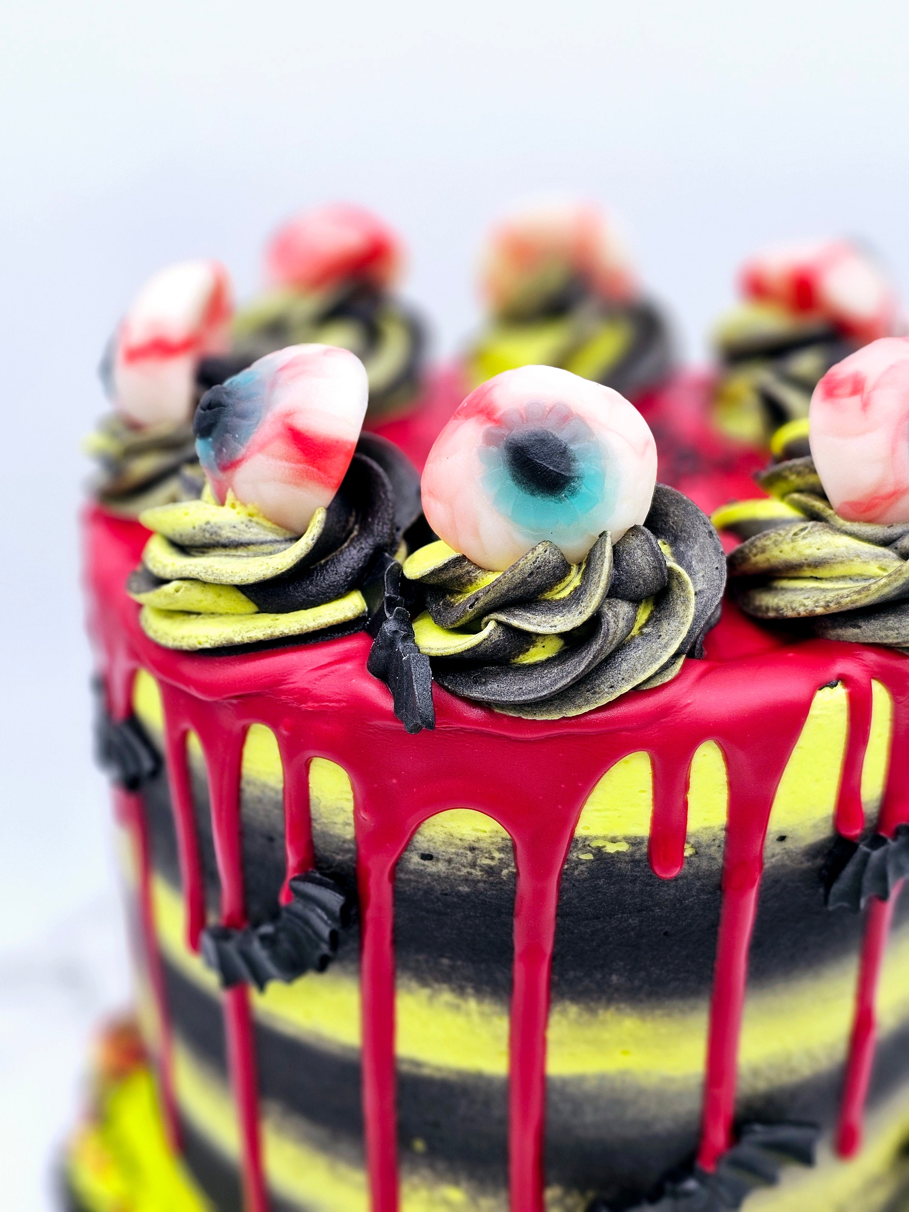 Close-up of a Halloween-themed cake by Cake Secret, featuring red drip icing, black and yellow buttercream swirls, and decorated with edible eyeballs on top, with spooky bat decorations, displayed on a marble-effect cake stand.