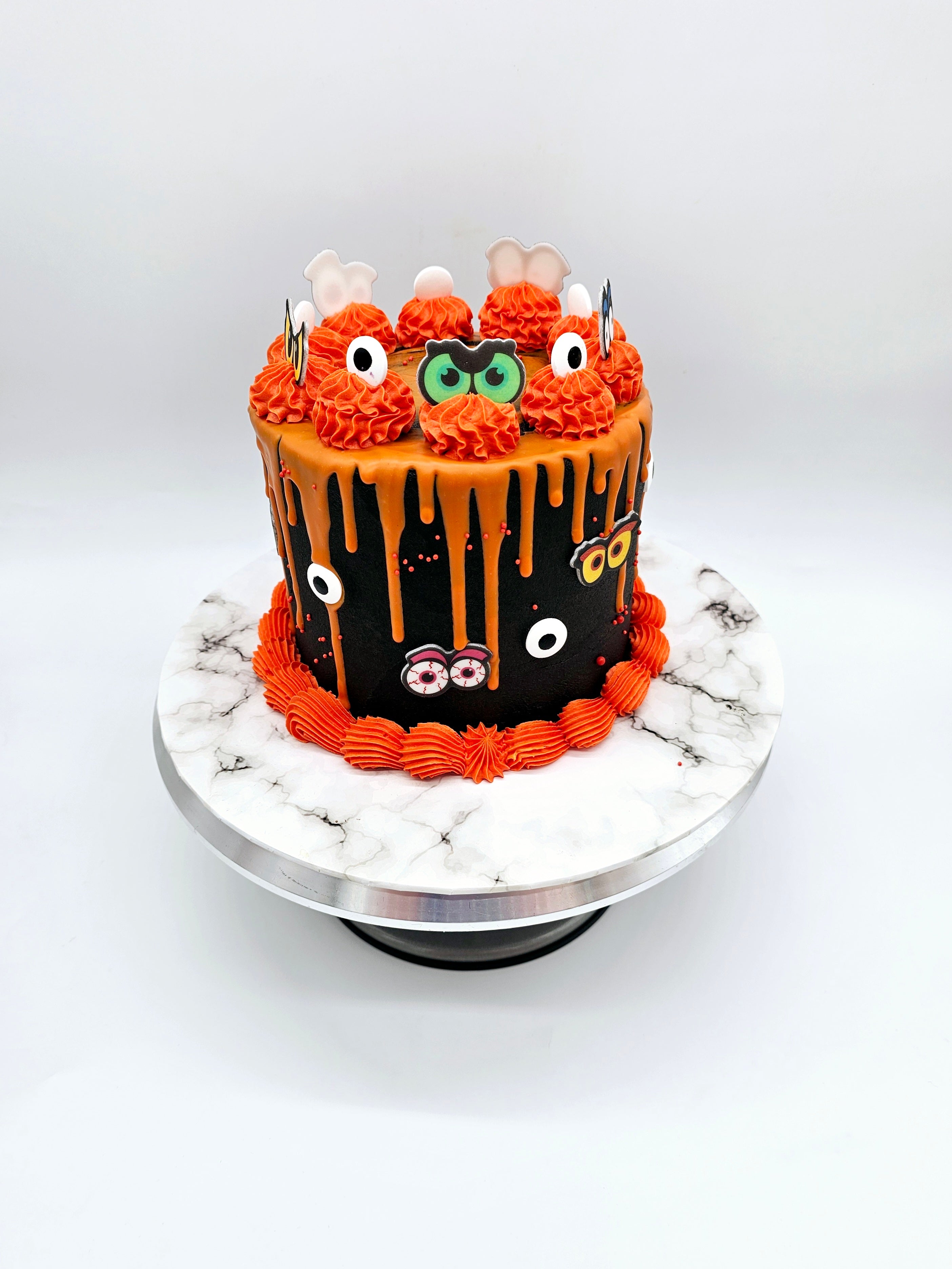 A spooky Halloween-themed cake by Cake Secret, featuring orange drip icing over a black buttercream base, decorated with piped orange swirls, fondant eyes, ghosts, and eyeball details, displayed on a marble-effect cake stand.