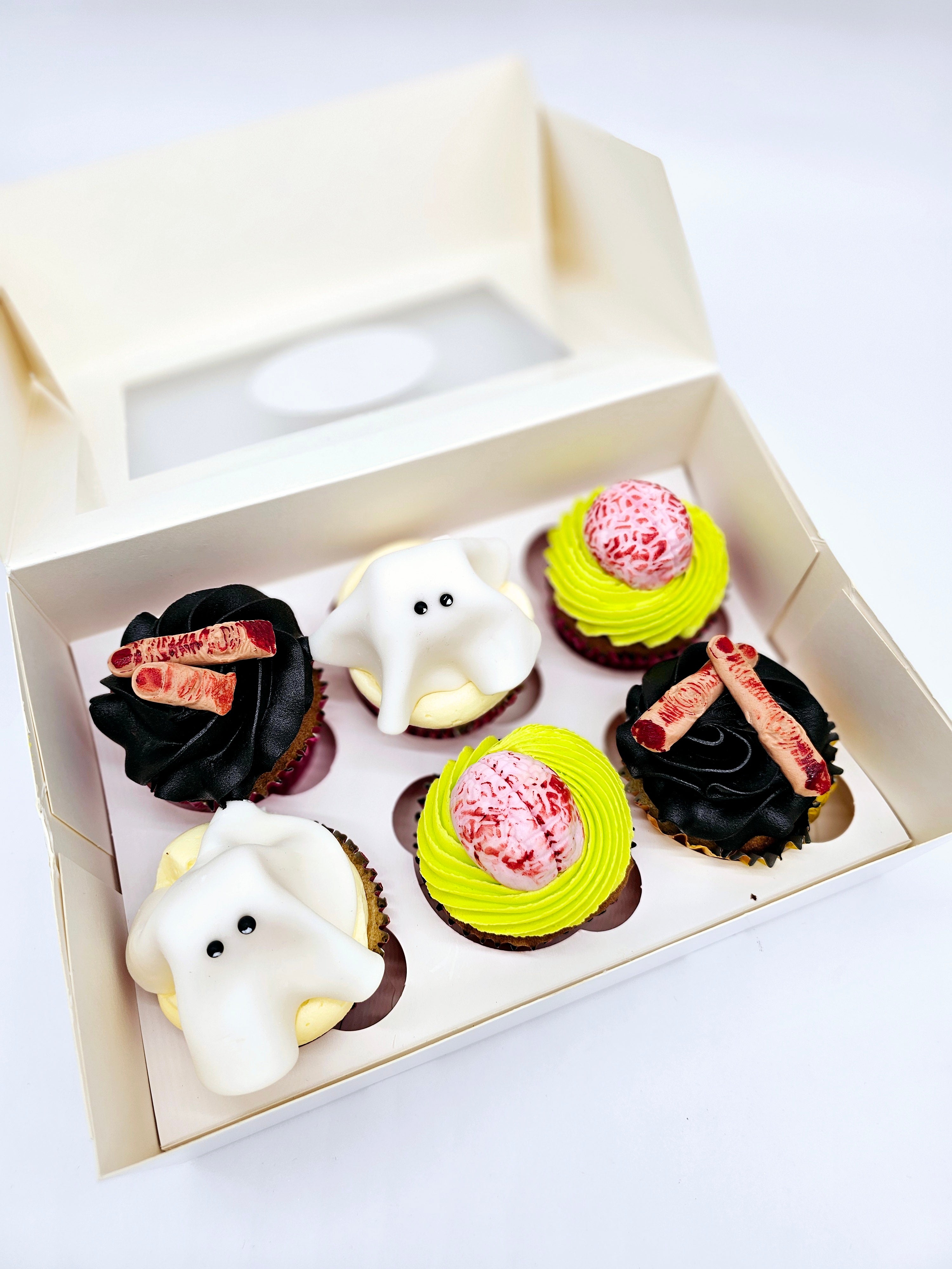 Top view of a box of six Halloween-themed cupcakes by Cake Secret, featuring ghost-shaped fondant toppers, edible brains, and severed finger decorations on black and neon green buttercream, displayed in an open cupcake box.