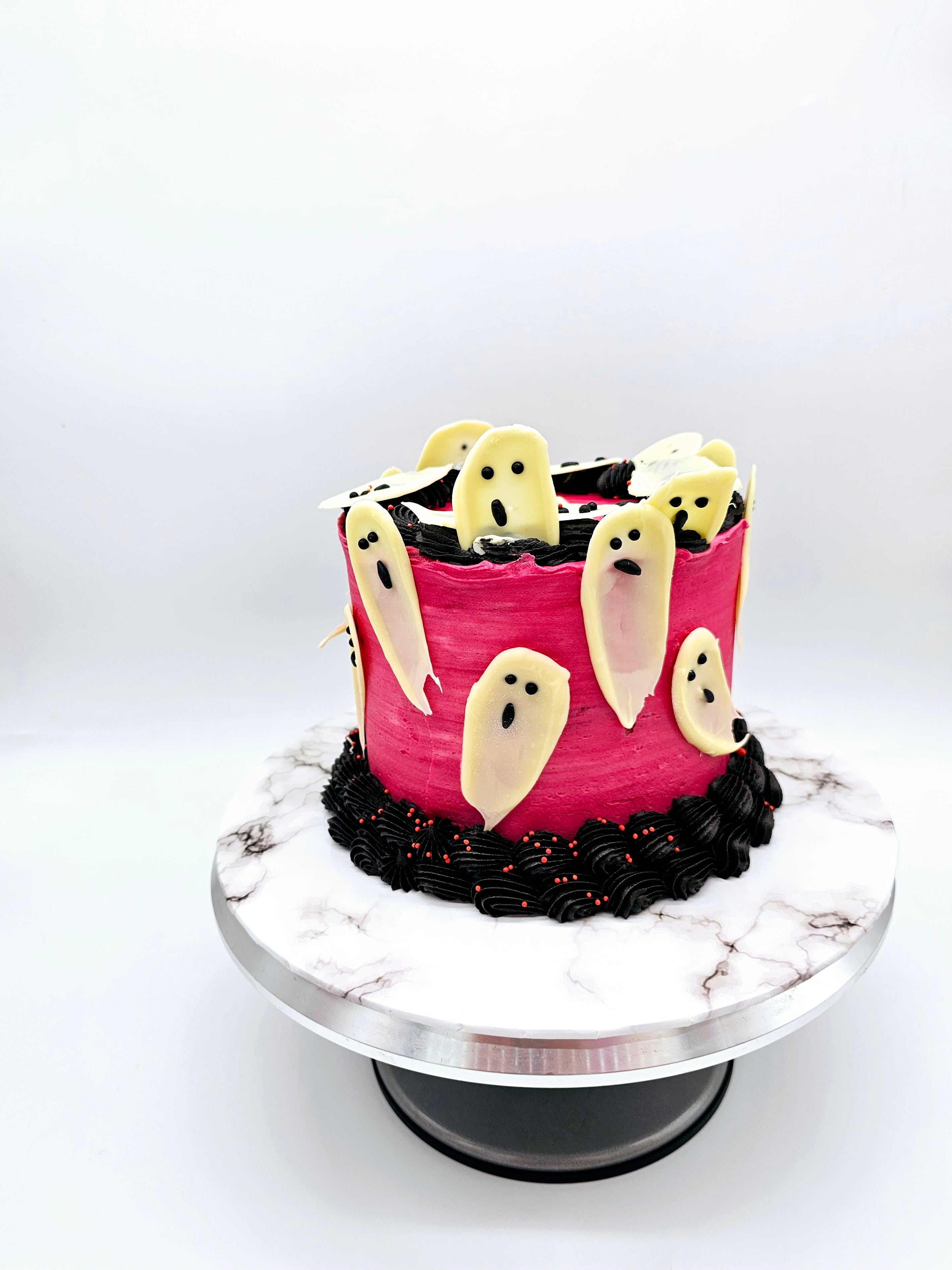 Side view of a Halloween-themed cake featuring white chocolate ghost decorations on a pink buttercream background, with black and red piped swirls along the base, created by Cake Secret.