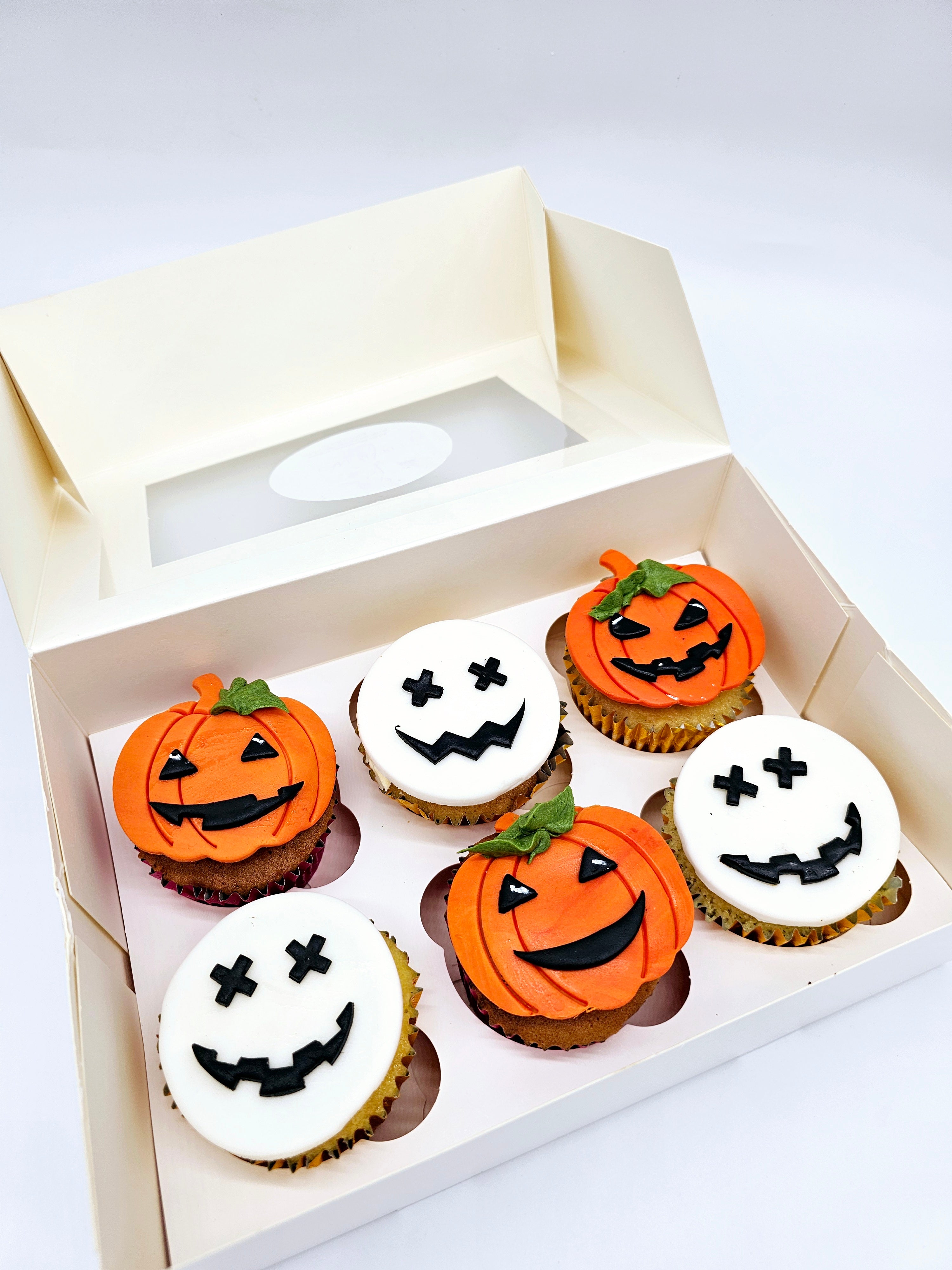 Close-up of a box of six Halloween-themed cupcakes by Cake Secret, decorated with orange pumpkin faces and white skull faces, each with various black icing expressions, displayed in an open cupcake box.