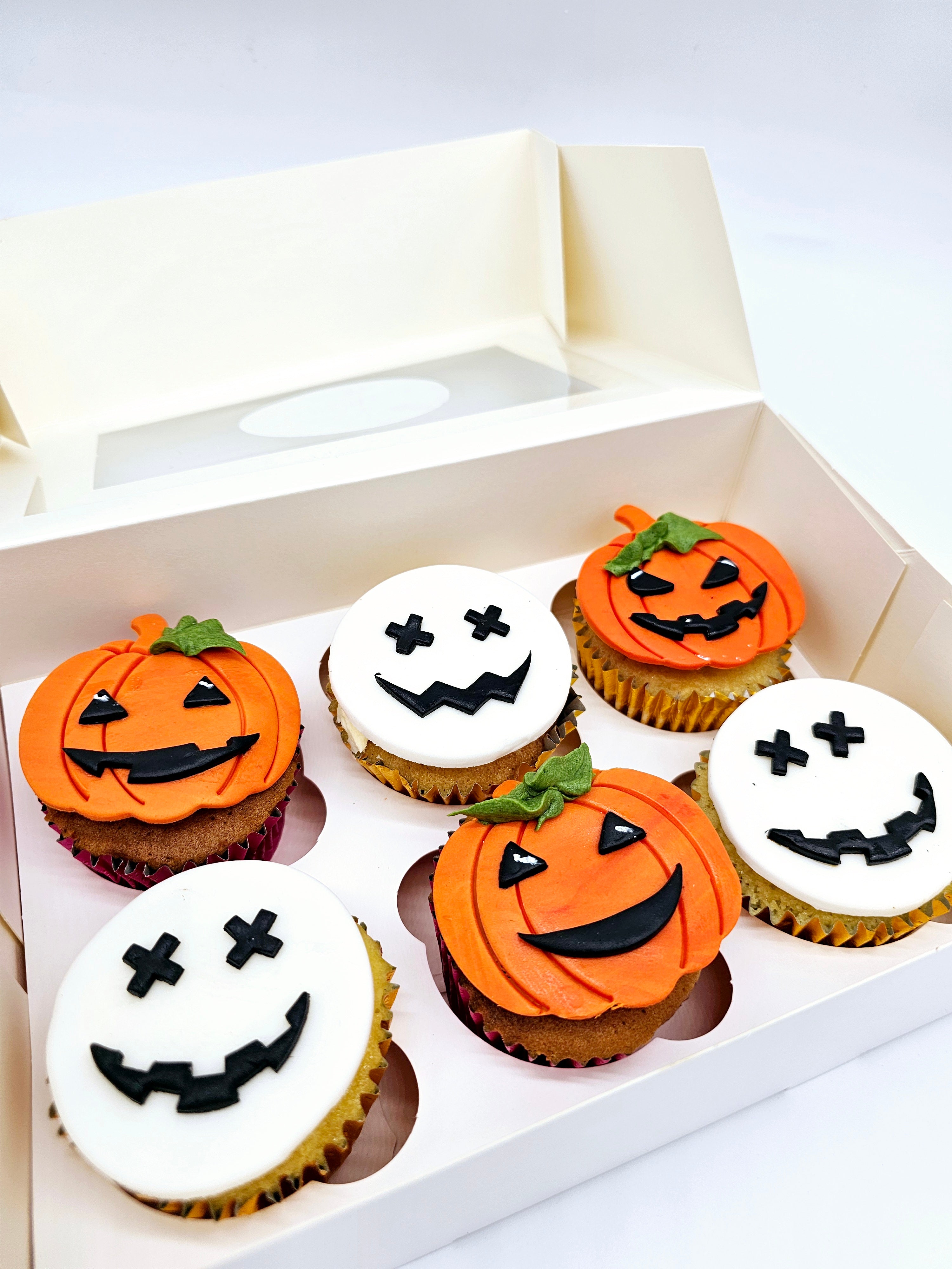 Close-up view of six Halloween-themed cupcakes by Cake Secret, featuring orange pumpkin faces and white skull faces with various black icing expressions, displayed in an open cupcake box.