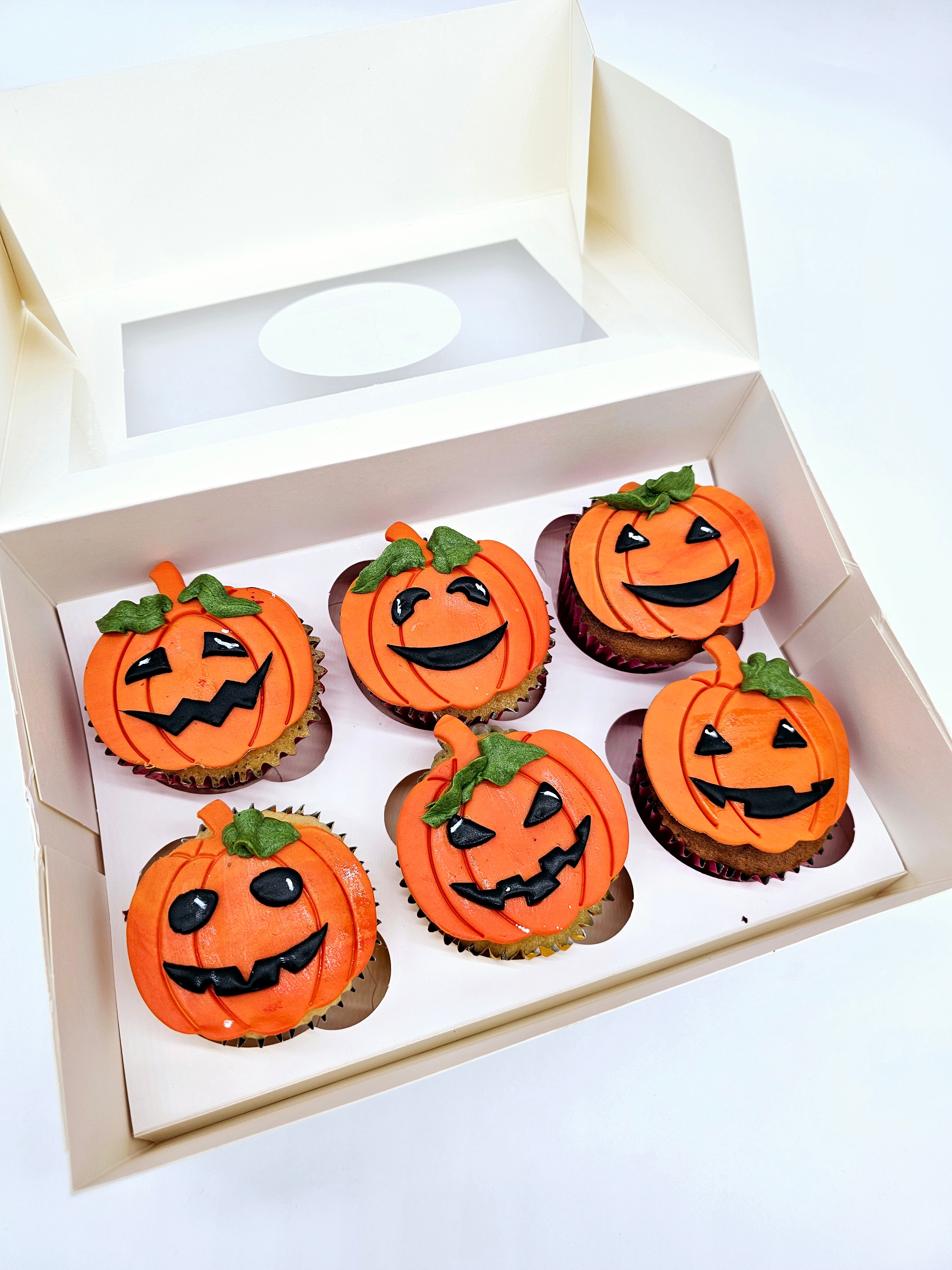 A box of six Halloween-themed cupcakes by Cake Secret, each decorated with orange pumpkin faces featuring various spooky and smiling expressions in black icing, presented in an open cupcake box.