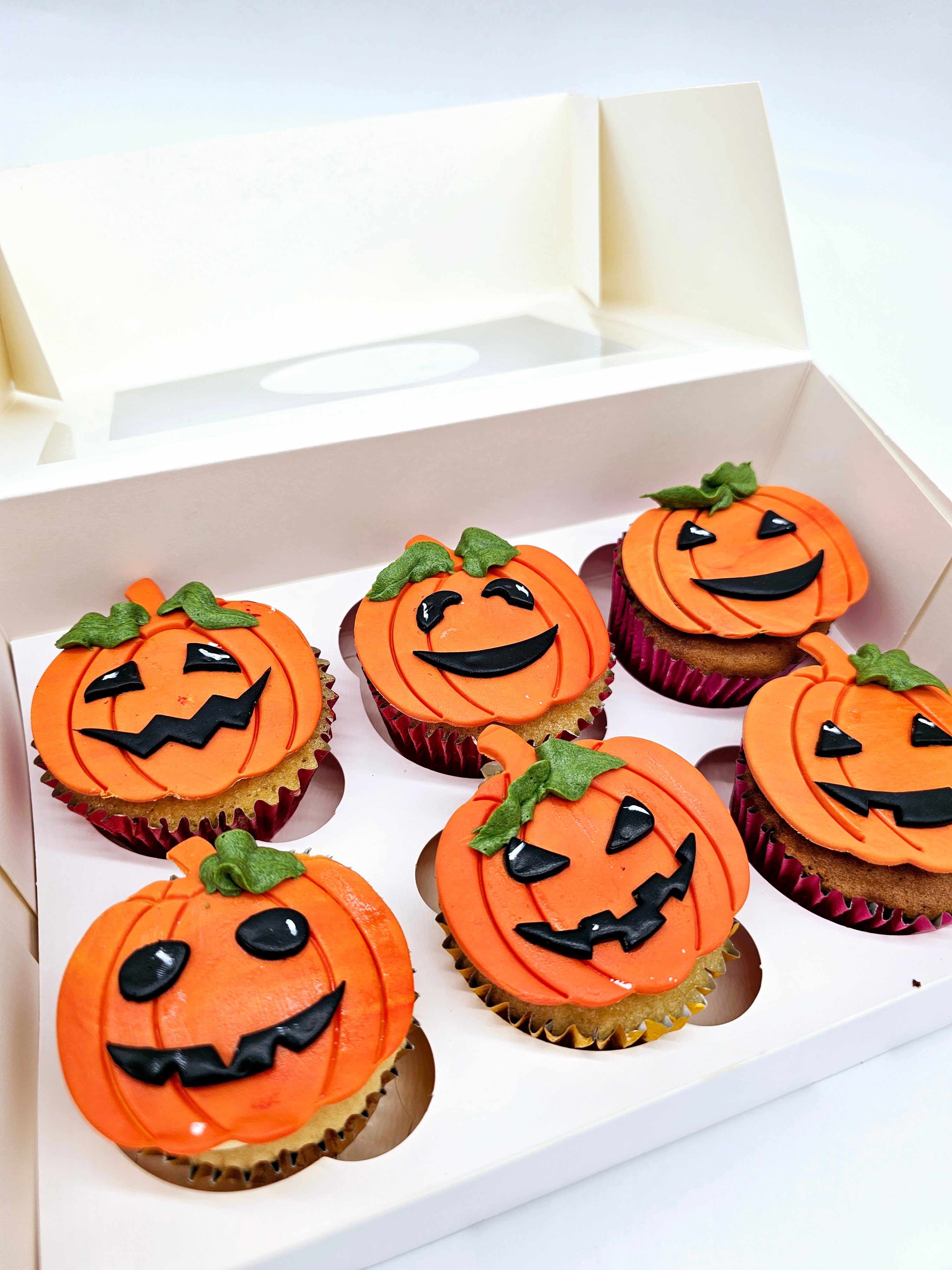 Close-up view of six Halloween-themed cupcakes by Cake Secret, each decorated with orange pumpkin faces featuring various spooky expressions in black icing, displayed in an open cupcake box.