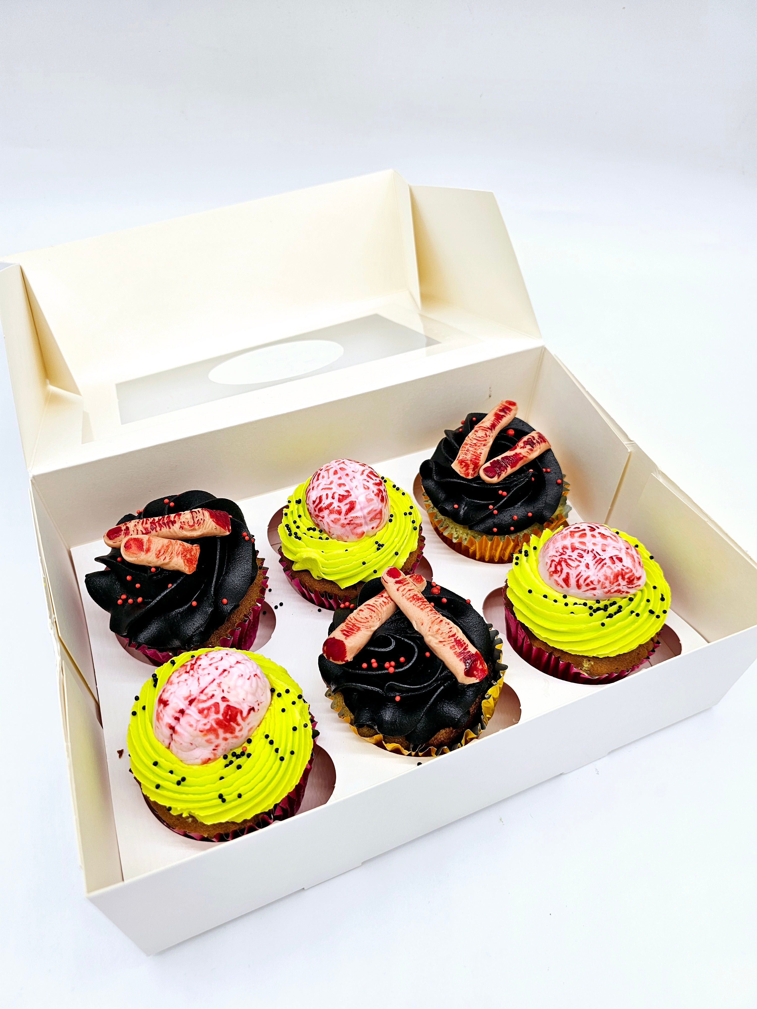 A box of six Halloween-themed cupcakes by Cake Secret, featuring spooky decorations like edible brains on bright green frosting and severed fingers on black buttercream, with red and black sprinkles.