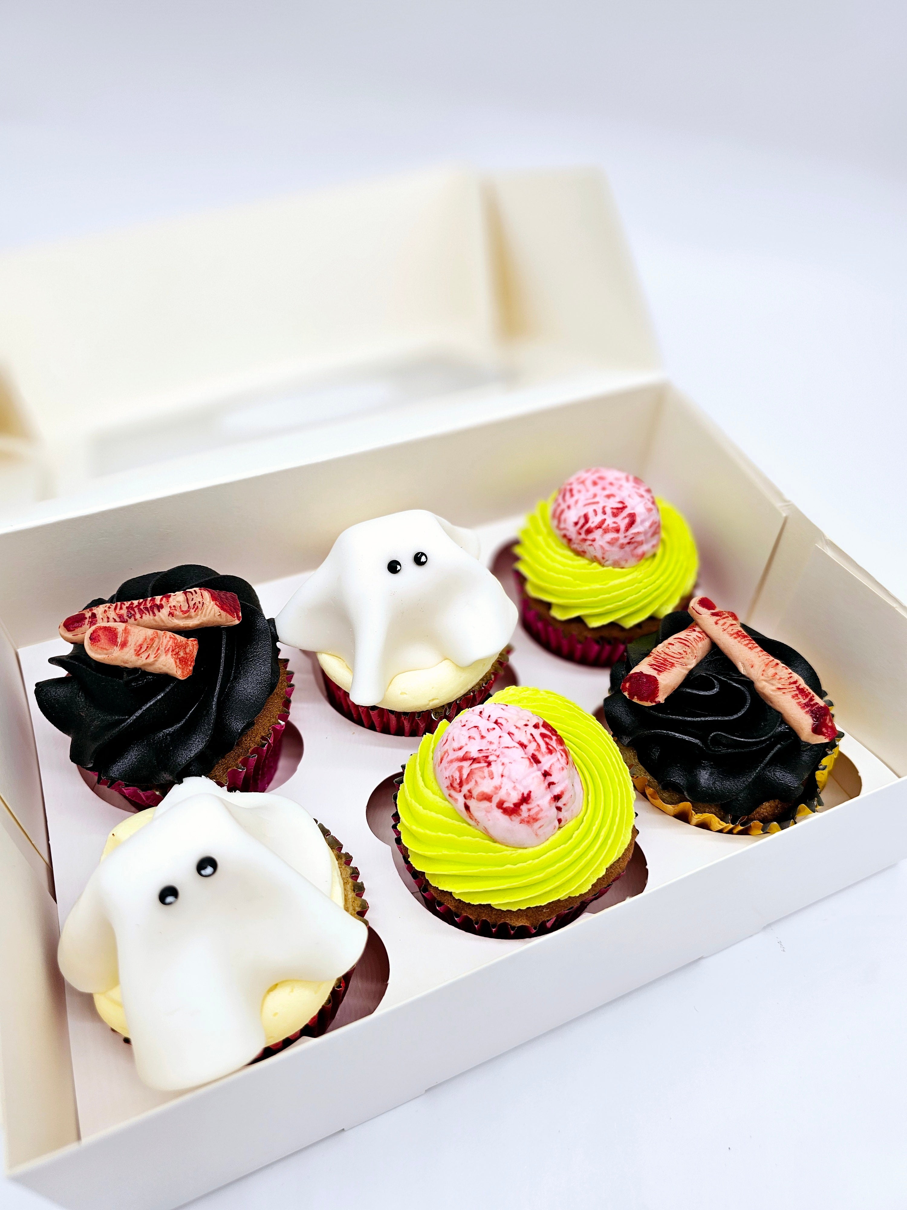 A box of six Halloween-themed cupcakes by Cake Secret, featuring ghost-shaped fondant toppers, edible brains, and severed finger decorations on black and neon green buttercream, neatly displayed in an open cupcake box.