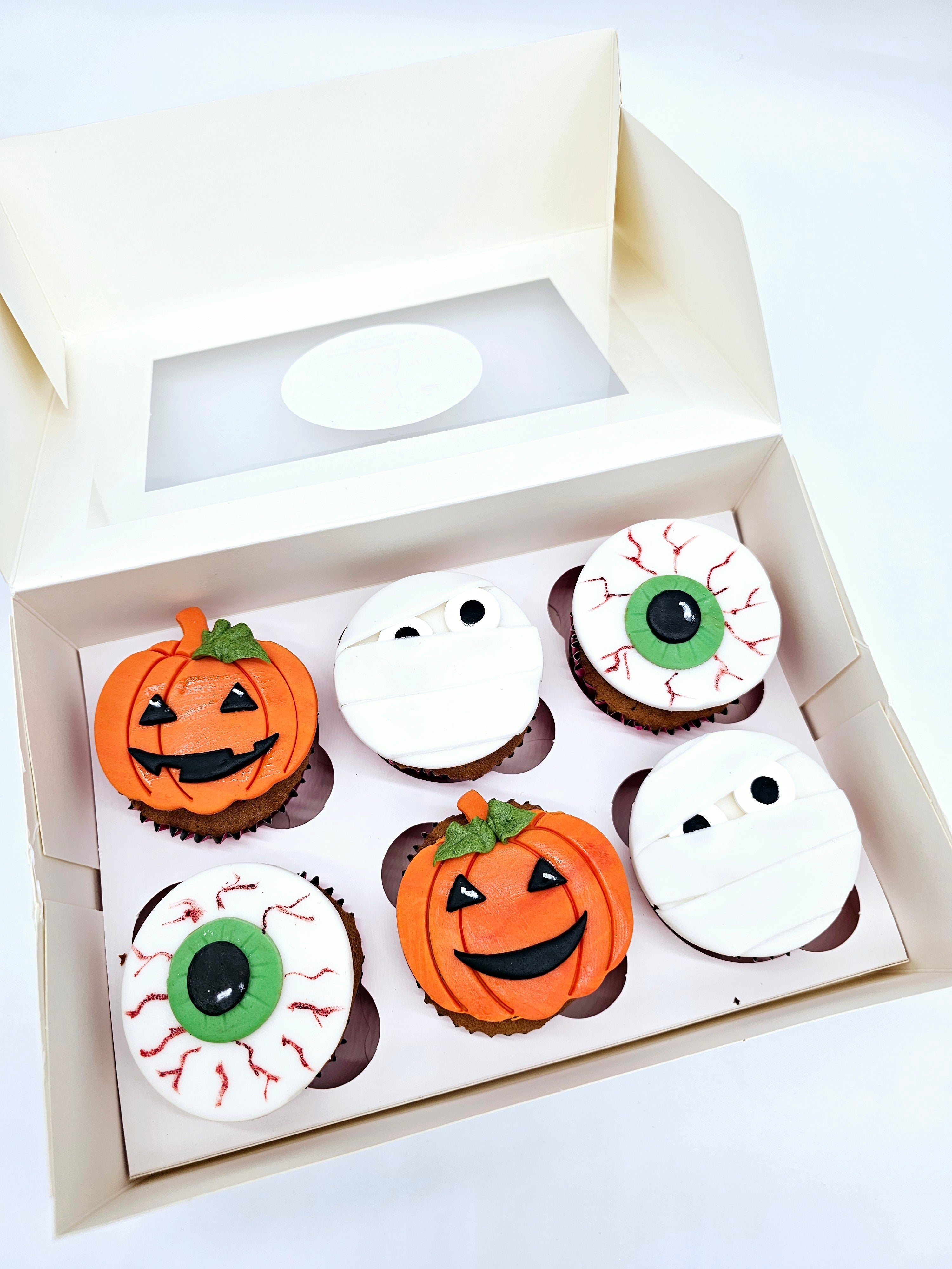 A box of six Halloween-themed cupcakes by Cake Secret, decorated with fondant toppers shaped as pumpkins, mummies, and eyeballs, neatly displayed in an open white cupcake box.