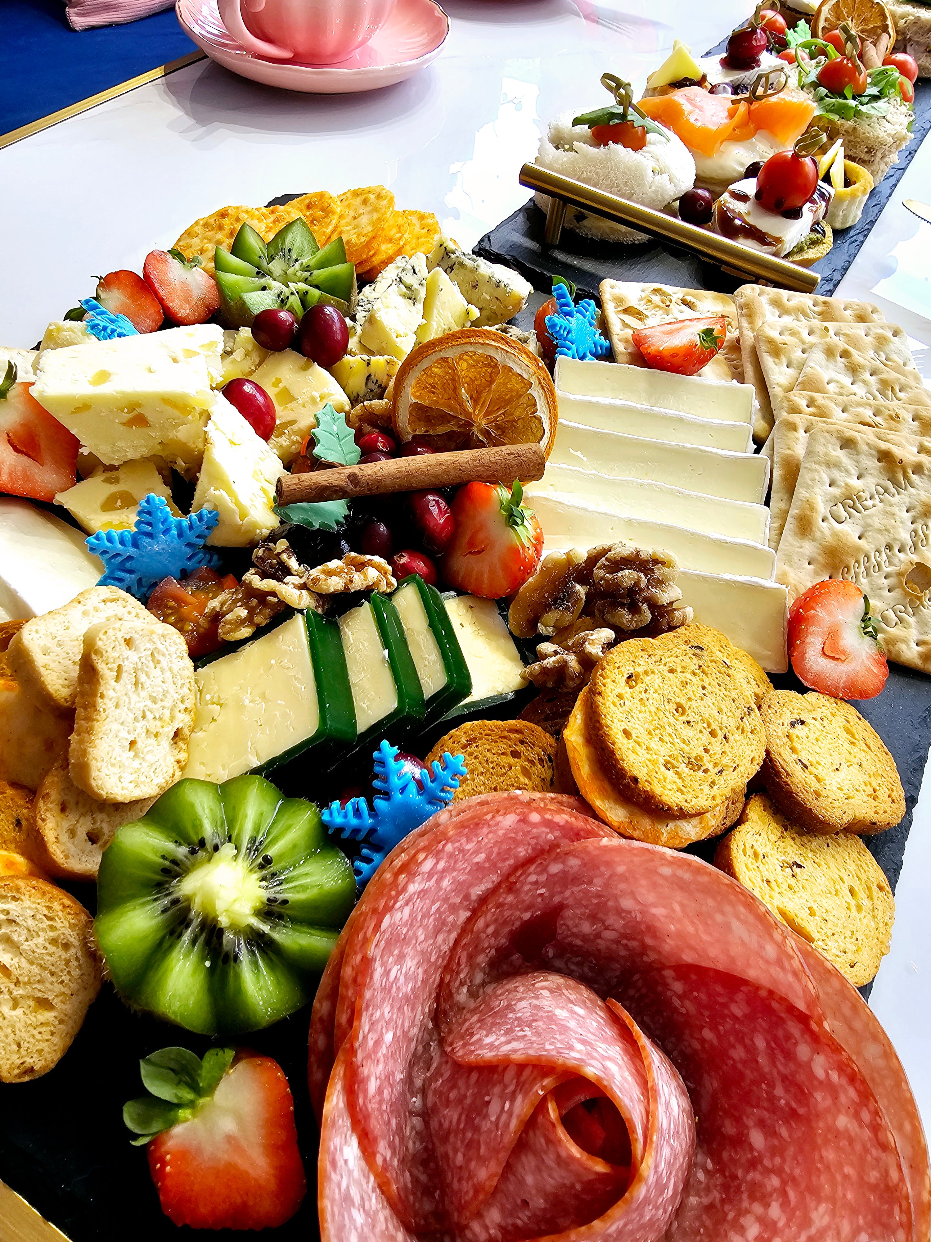 Wide view of a Christmas-themed cheese and charcuterie board from Cake Secret’s afternoon tea, featuring a variety of cheeses, salami, crackers, fresh fruit like strawberries and kiwi, walnuts, cranberries, dried orange slices, and a cinnamon stick, with blue snowflake decorations.