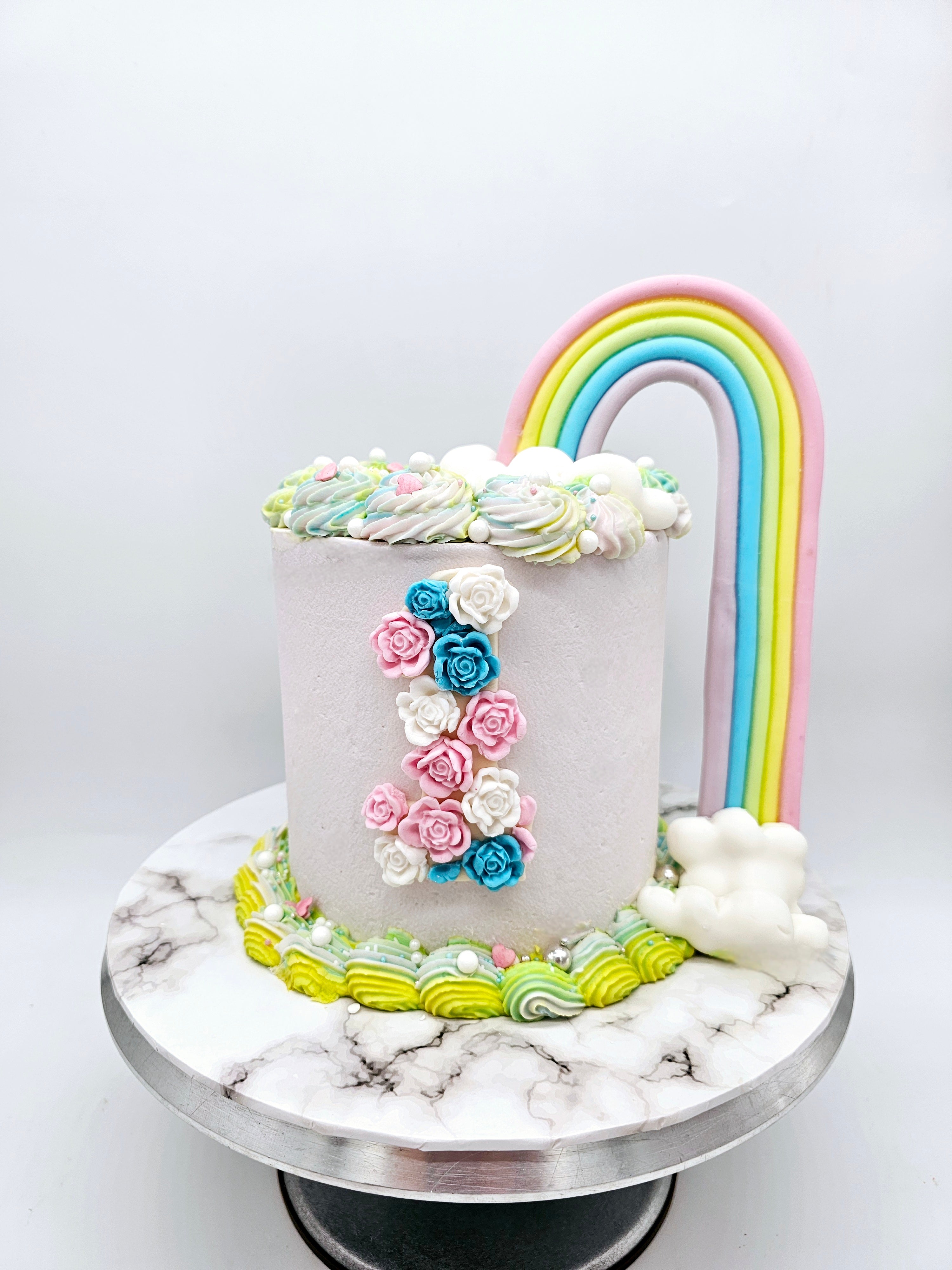 A pastel-coloured first birthday cake made by Cake Secret, featuring a fondant rainbow arch and floral details forming the number one, displayed on a marble-effect cake stand.