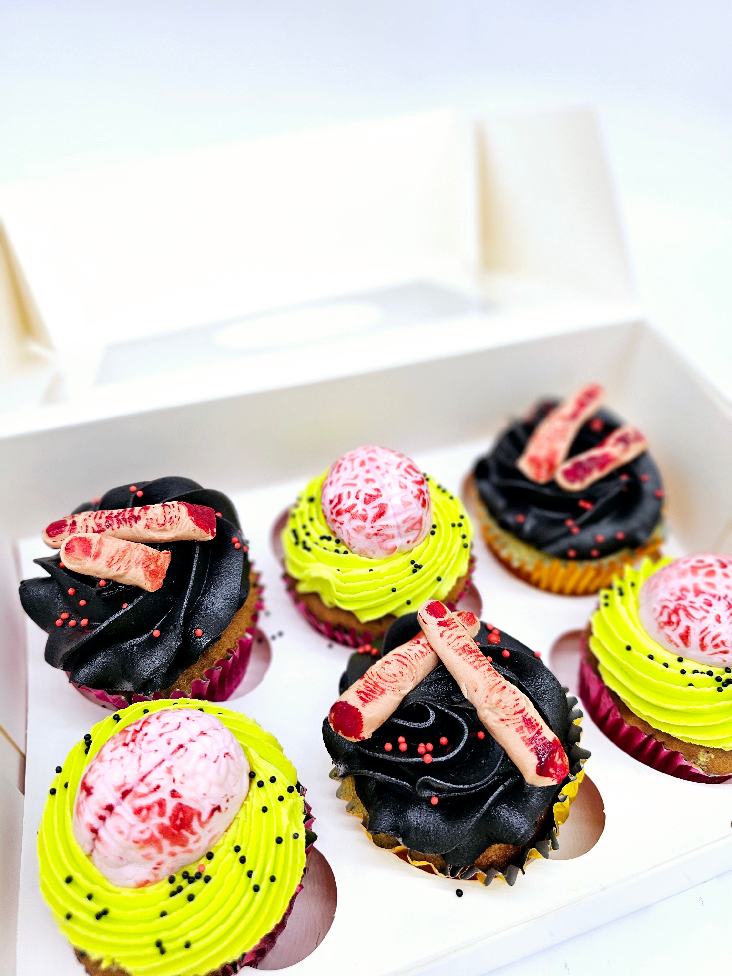 Box of six Halloween-themed cupcakes featuring edible brains and severed fingers, with neon green and black frosting, and creepy sprinkle decorations.