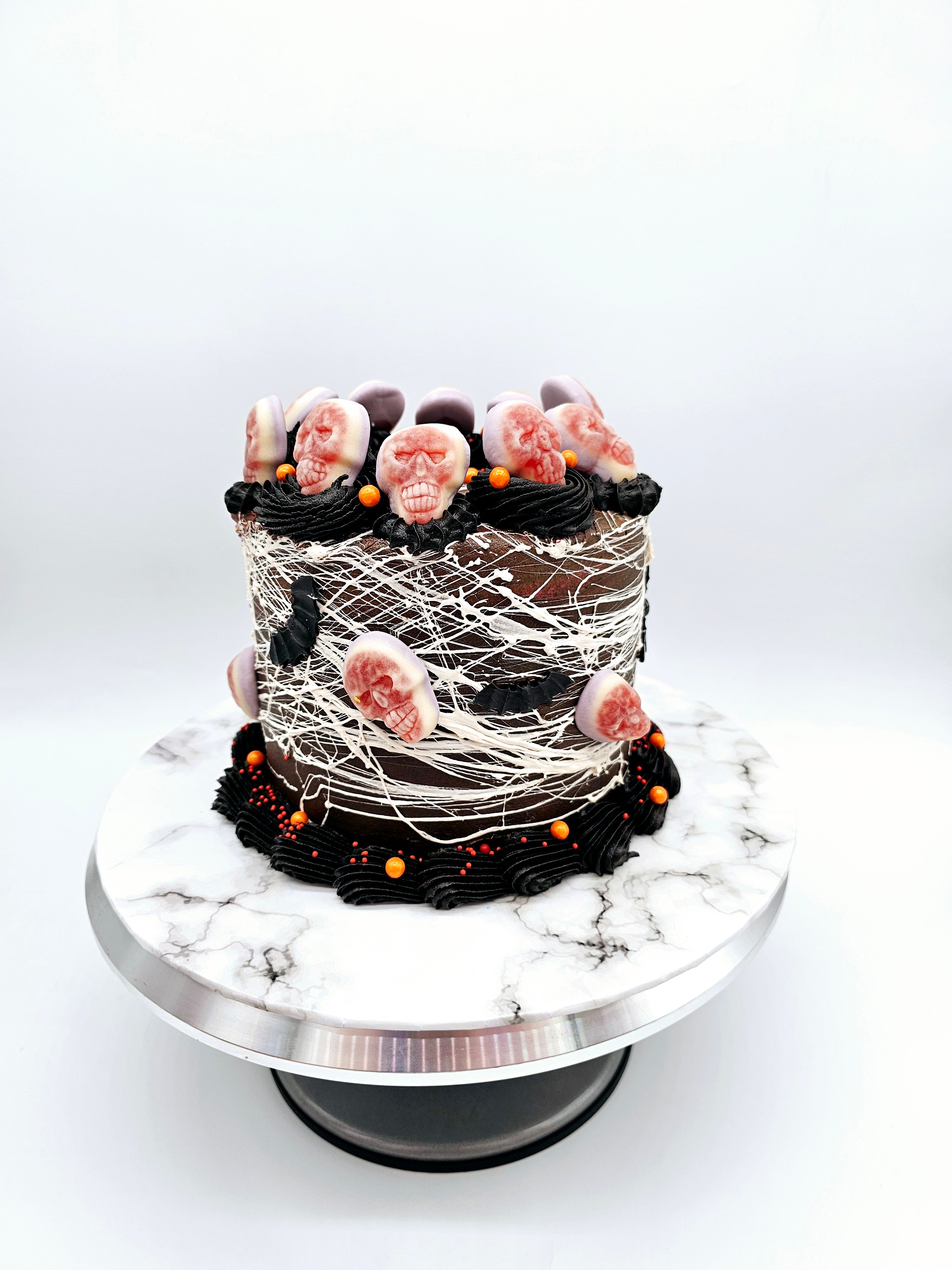 Side view of a Halloween-themed cake wrapped in white spider-web-like strands, topped with edible skulls, black frosting, and orange sprinkle accents.