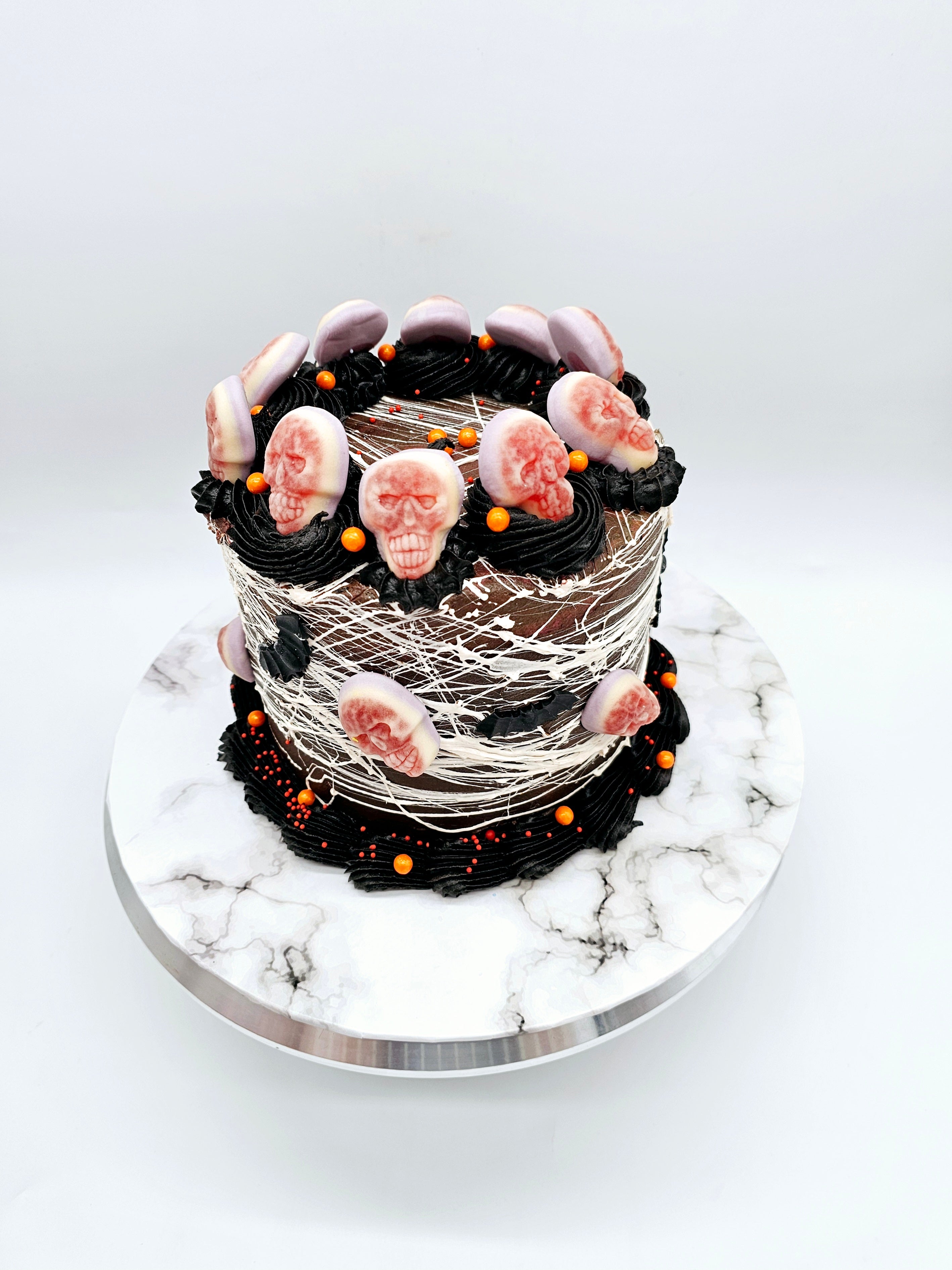 Halloween cake wrapped in white spider-web-like strands, decorated with skull toppers, black frosting, and orange sprinkle accents.